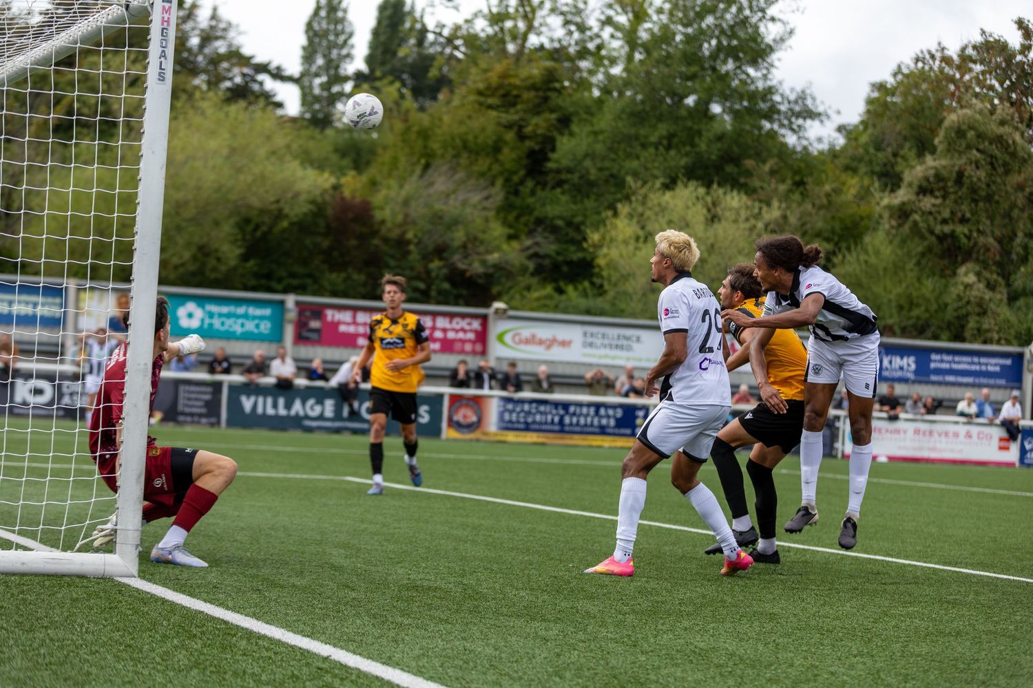 Soccer players compete in a header duel near the goal during an outdoor match on a green pitch.