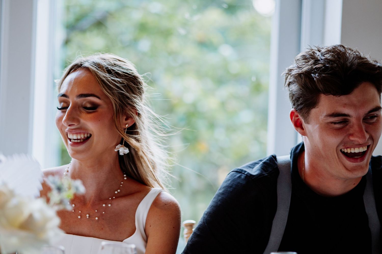 Bride and groom laughing joyfully at wedding reception table with floral centerpiece.