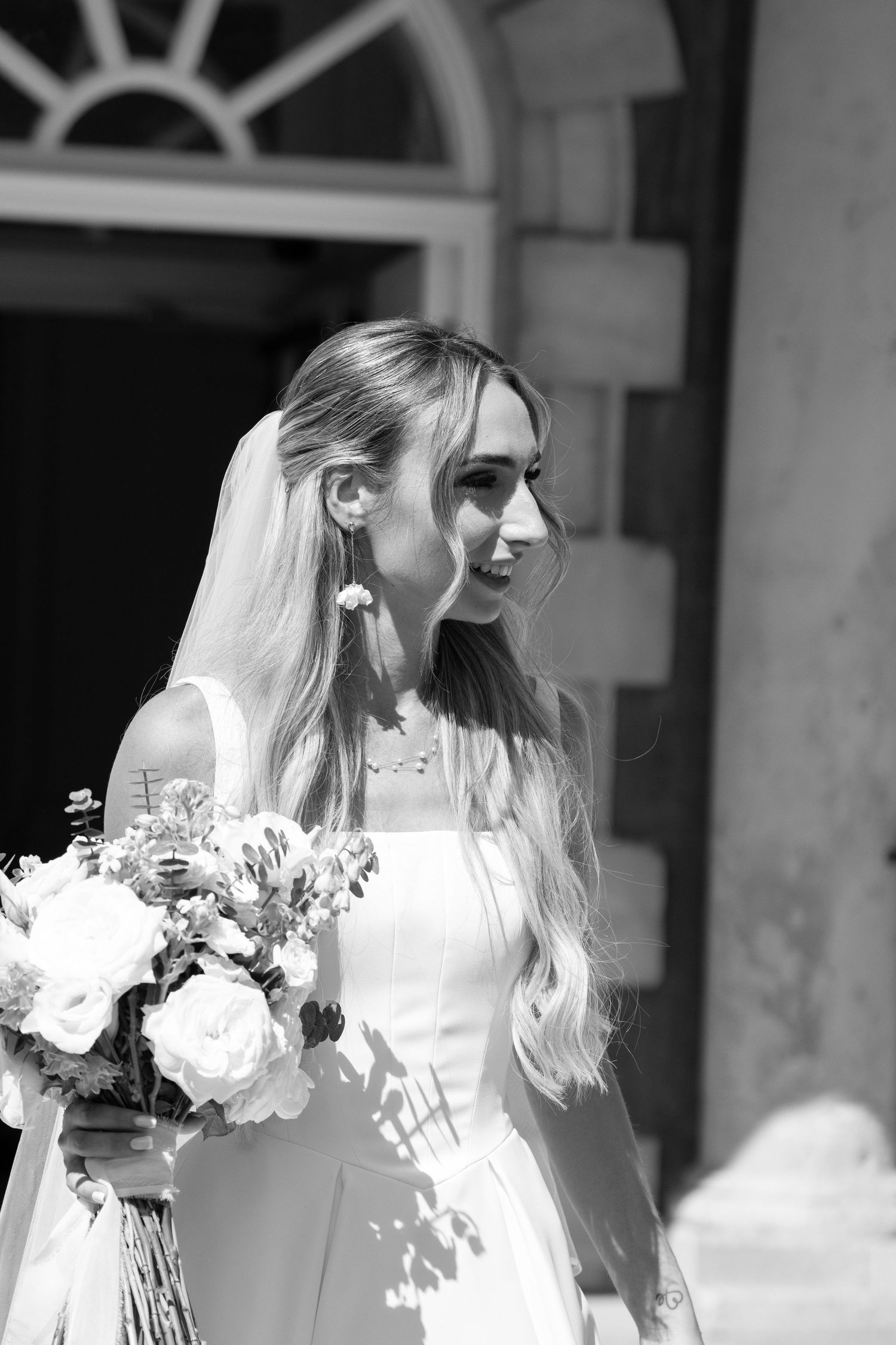Black and white portrait of elegant bride with long blonde hair holding white floral bouquet in sunlight.