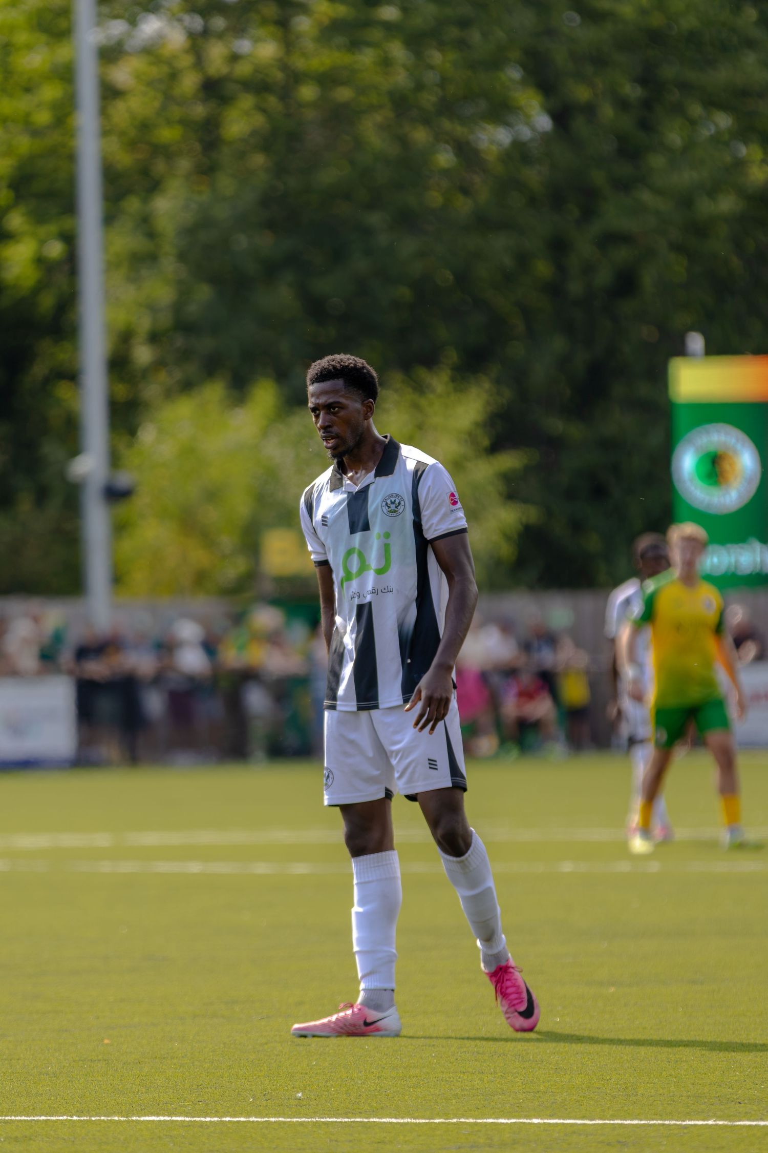 A footballer in black and white striped kit stands on a sunny green pitch during a competitive football match.