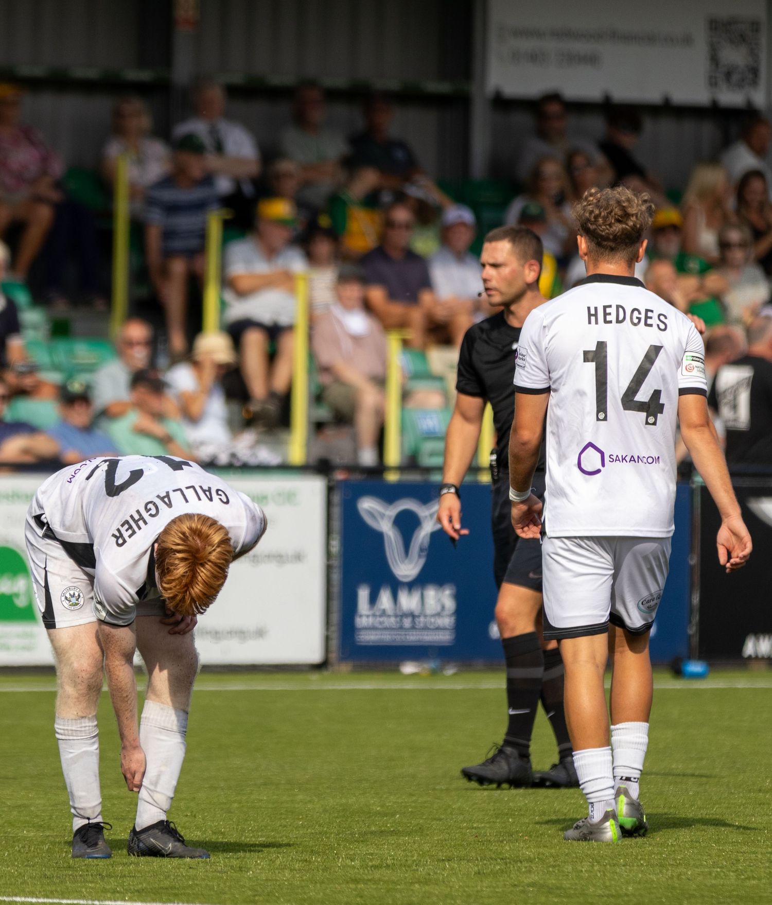 Gallagher bends down while Hedges #14 stands near a referee during a pause in a non-league football match.