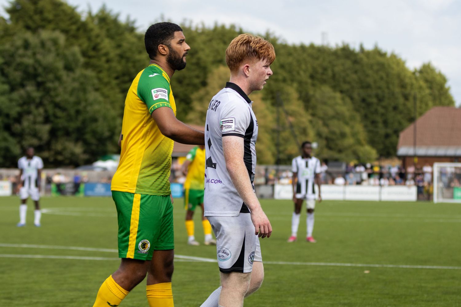 Football player in yellow kit gestures toward white-shirted number 2 opponent during outdoor match.