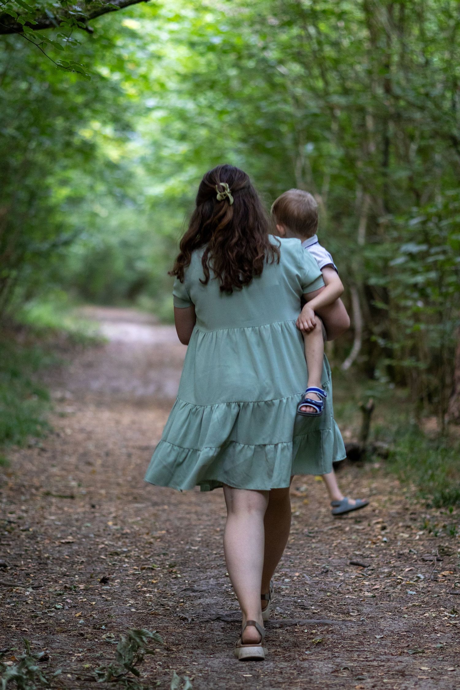 A woman in a sage green dress carries a toddler while walking along a forest path.