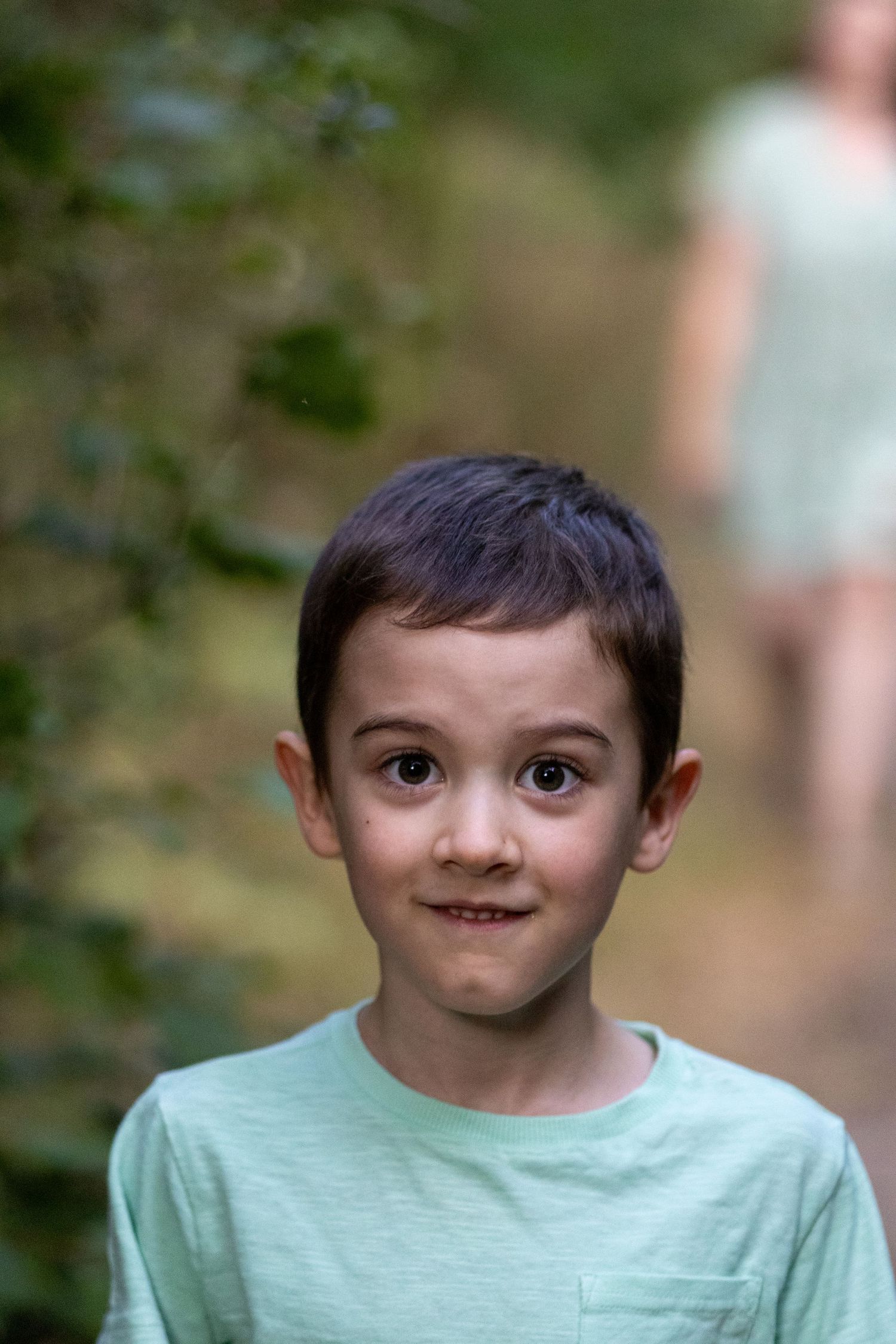 A dark-haired boy in a mint green t-shirt makes a funny face on a green forest path.