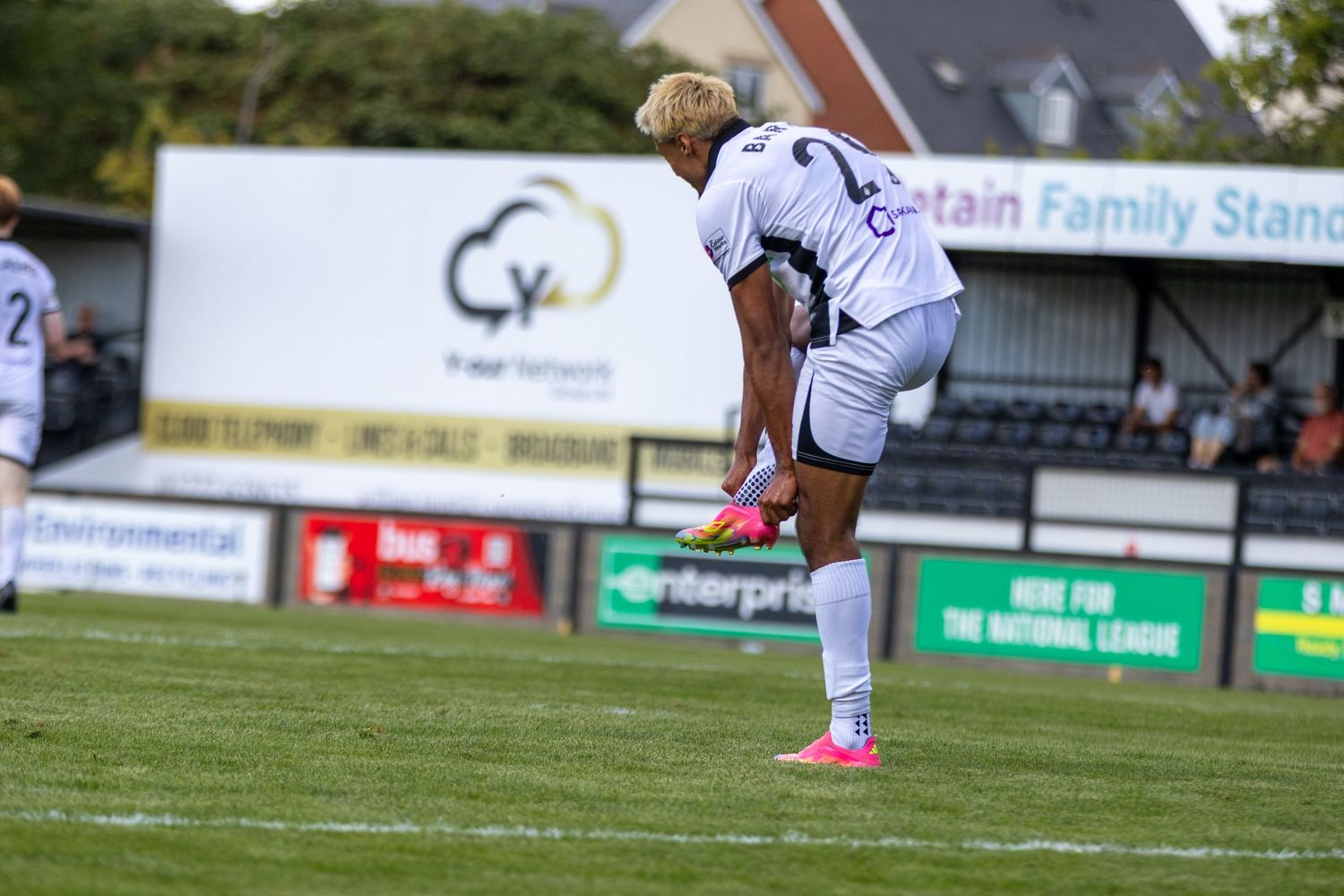 A white-kitted footballer performs a stretch near the Retain Family Stand during a non-league match.