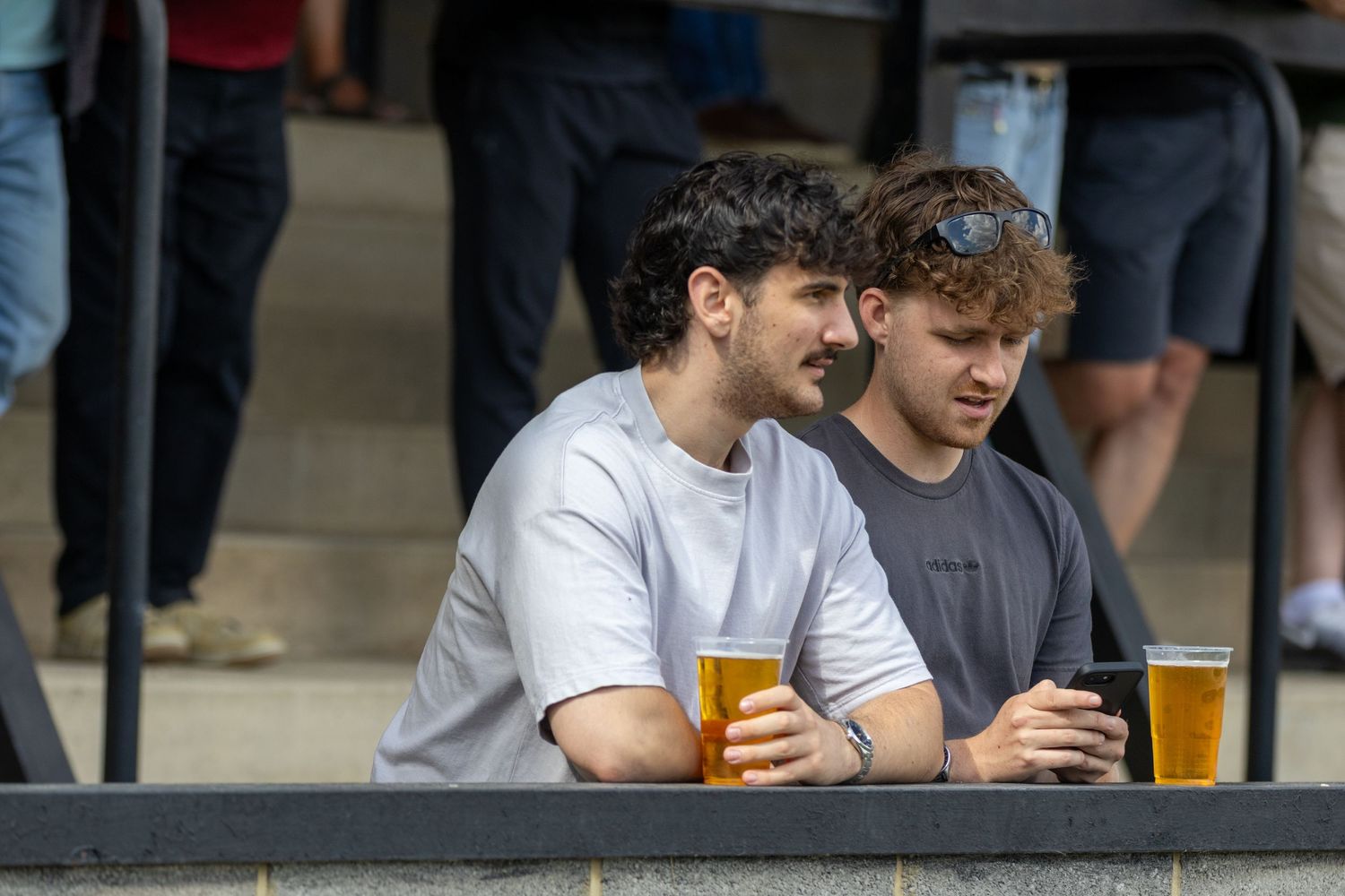 Two young men sitting outdoors holding pints of beer while one checks his smartphone.