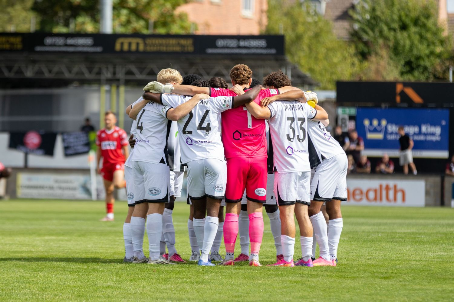 White-kitted football team huddle together on green pitch with pink goalkeeper in center before match.