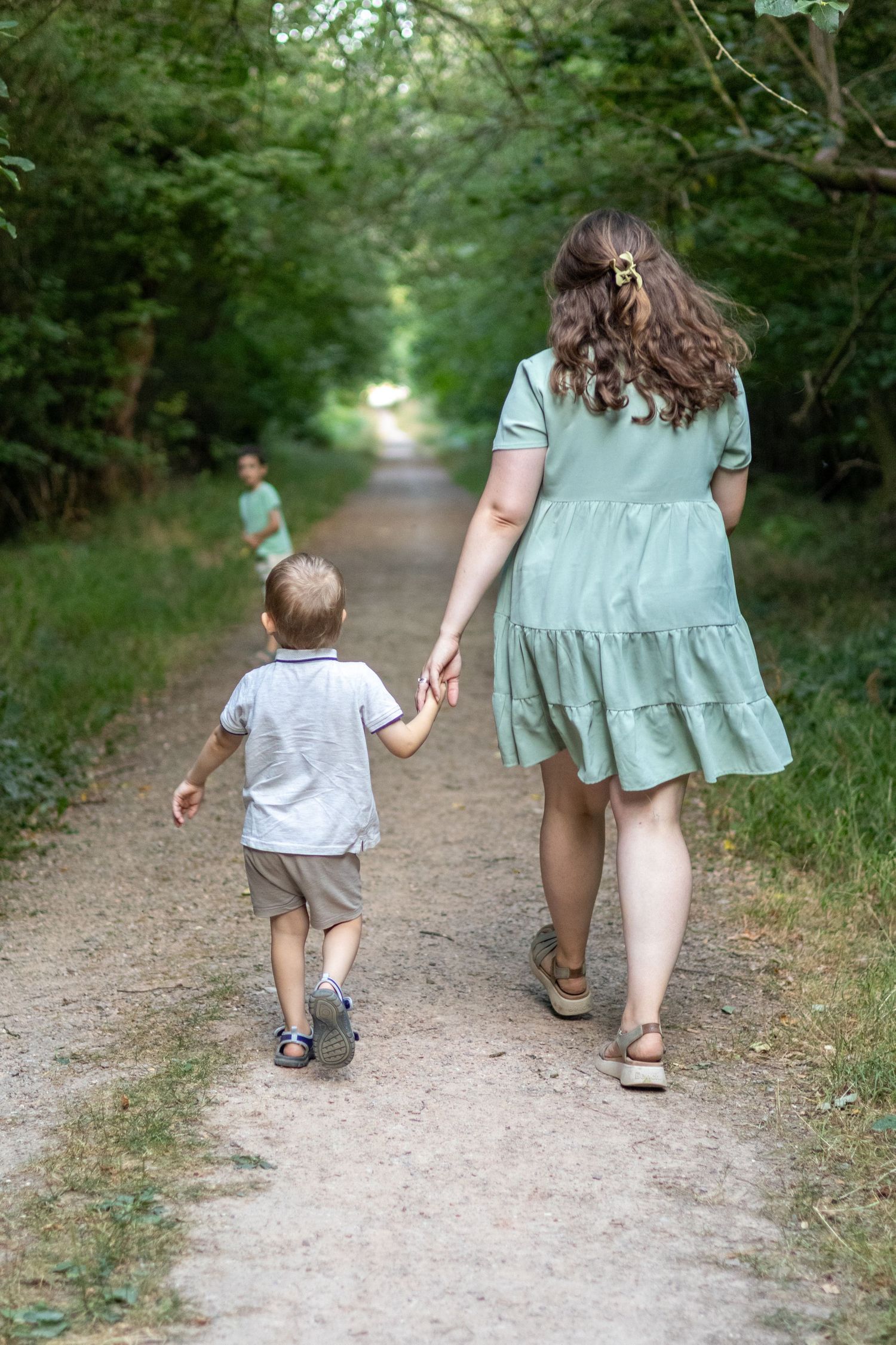 A woman in a green dress walks hand-in-hand with a young boy on a shaded woodland trail.