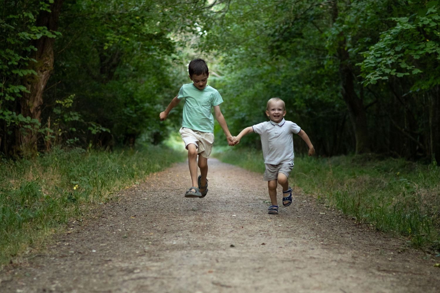 Two boys run hand-in-hand toward the camera on a lush green forest trail.