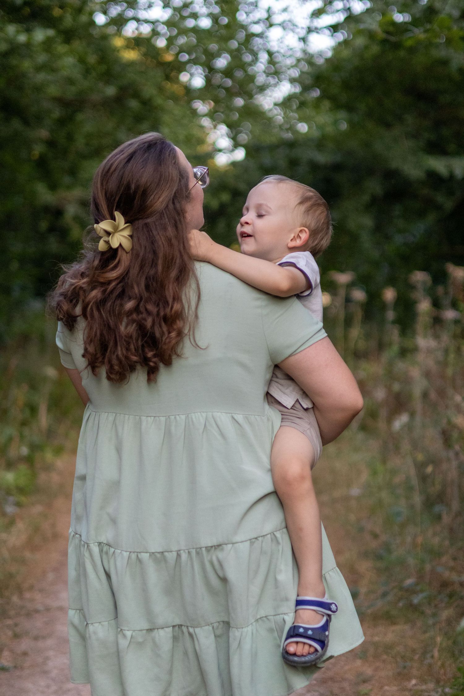 Woman in green tiered dress carrying a happy baby along a leafy outdoor pathway.