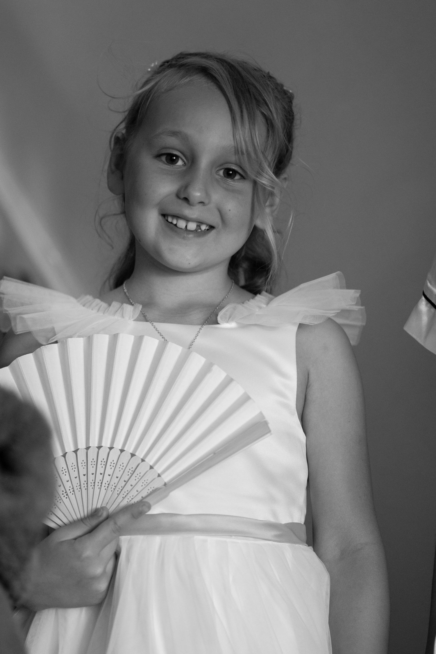 Young girl in white dress holding decorative fan smiles in black and white portrait photo.