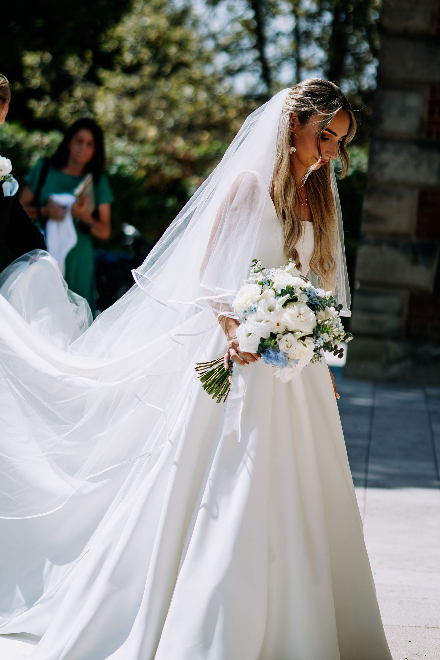 Stunning bride in white gown with billowing veil carrying white and blue flower bouquet.