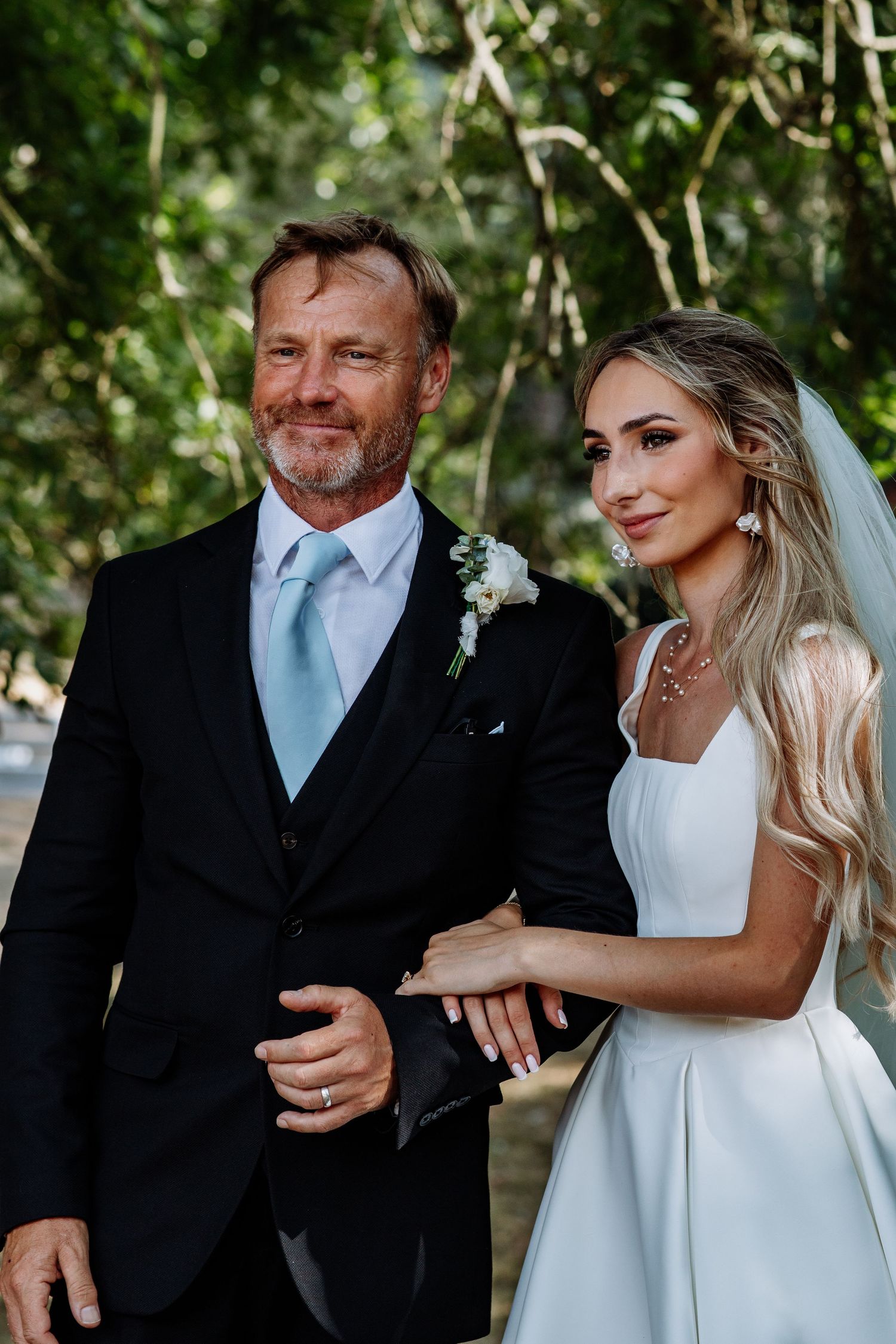 Elegant bride in white gown walking arm in arm with older gentleman in black suit at wedding.