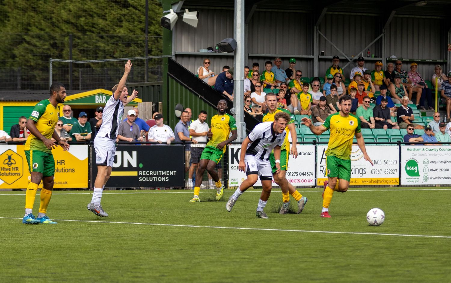 Football players in yellow-green and black-white kits compete during an outdoor match with spectators in the background.