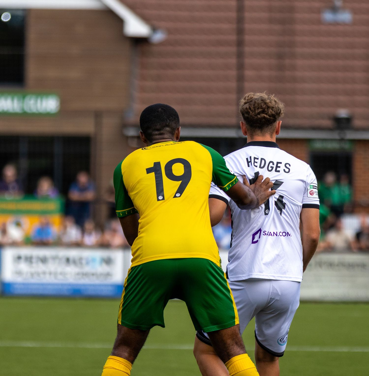 Number 19 yellow-kitted player battles for possession with Hedges in white during a football match.