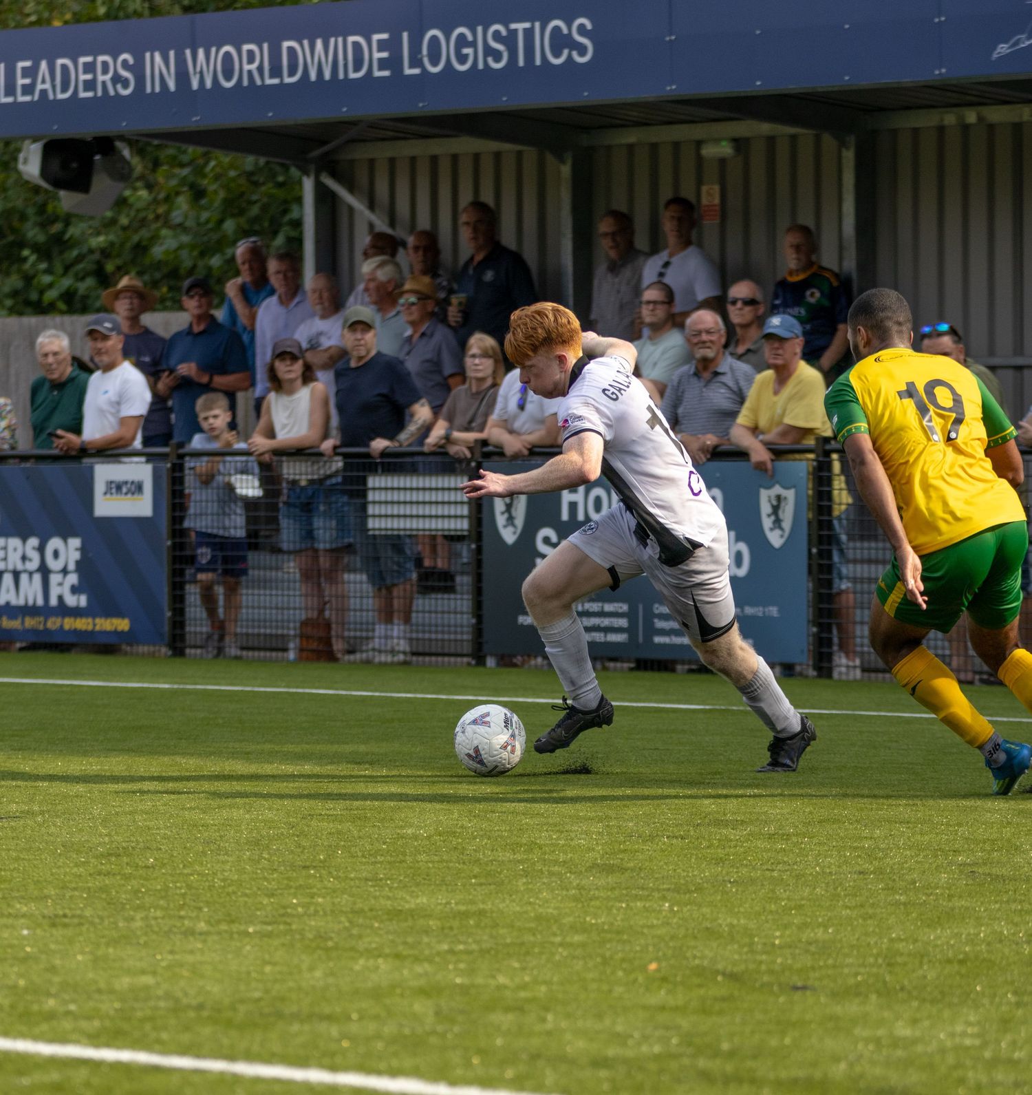 White-kitted footballer controls ball under pressure from number 19 opponent at Horsham FC stadium.