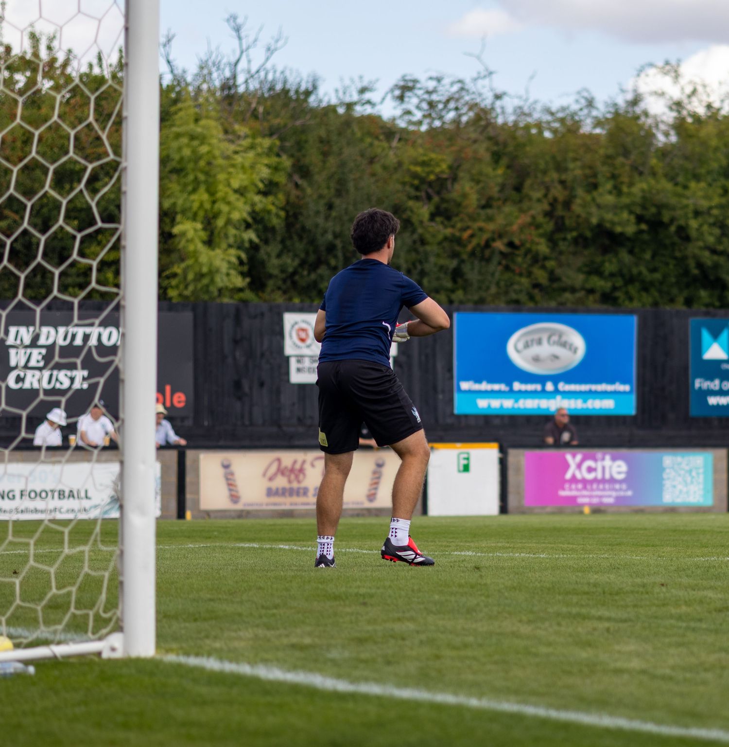 A football coach in navy kit stands by the goalpost observing play at a non-league football ground.