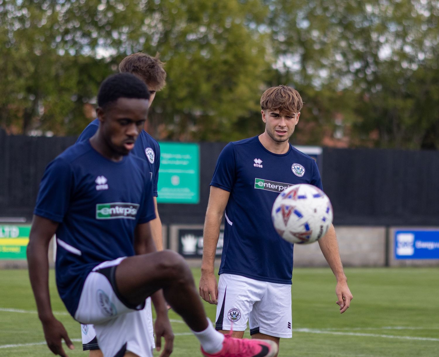 Two footballers in navy blue kits practice ball control during a pre-match warmup session on a grass pitch.