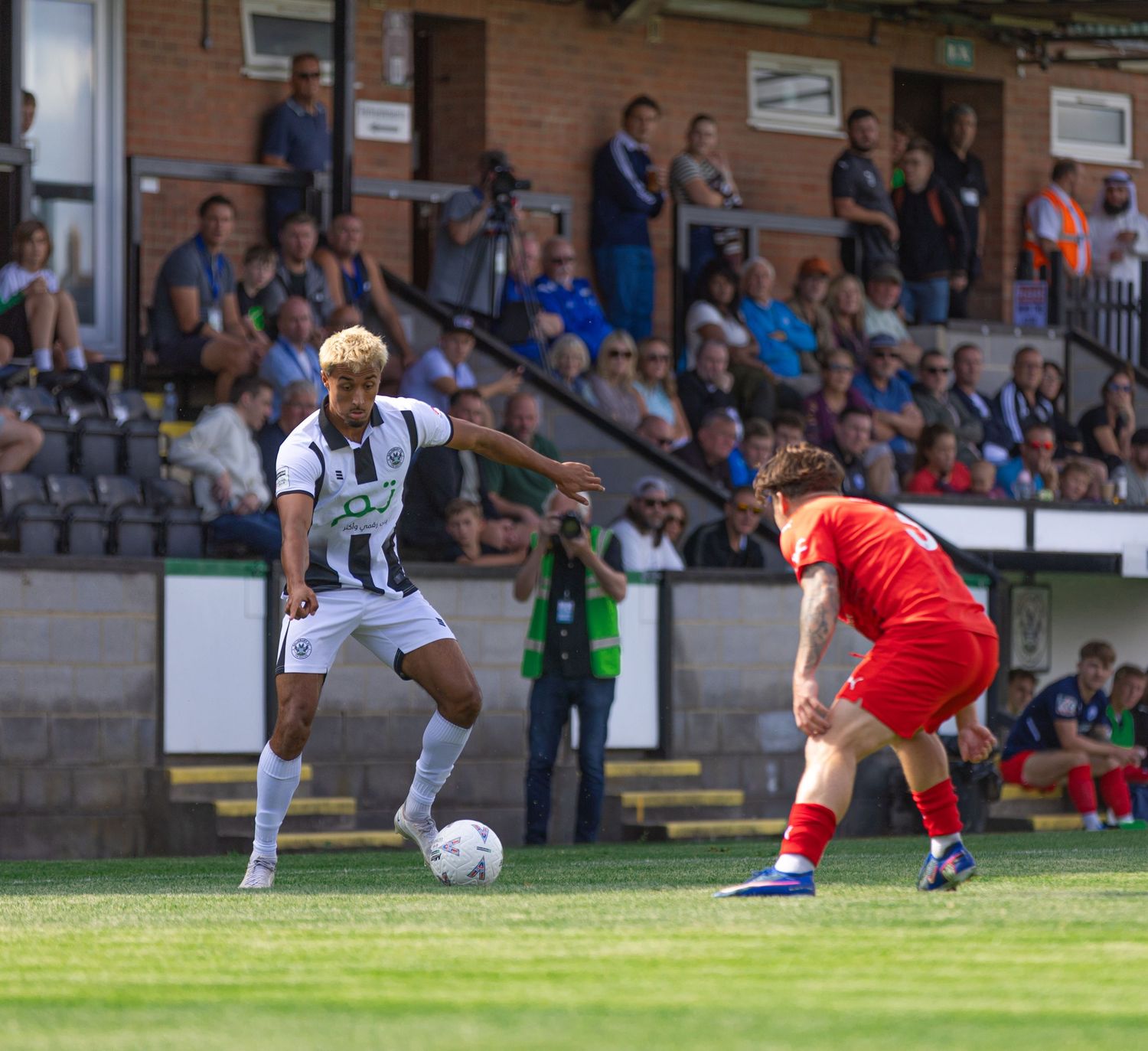 Two soccer players in black-and-white and red kits compete for the ball on a sunny green pitch with spectators watching.