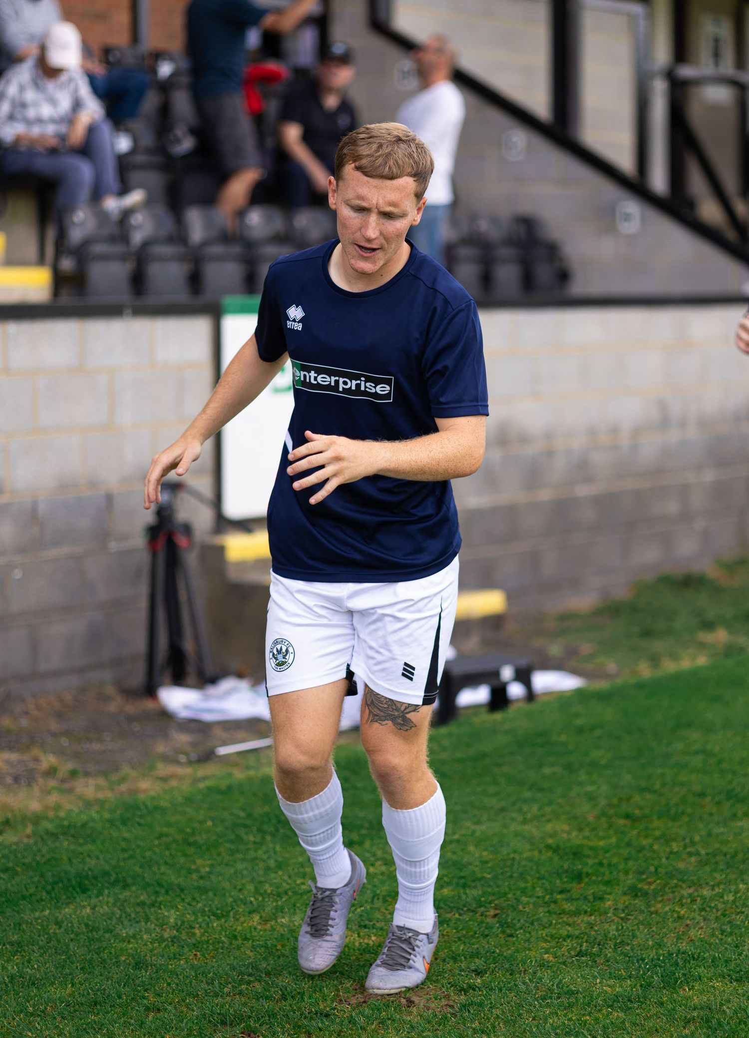 A male soccer player in navy blue jersey and white shorts walks on a grass field during a match.