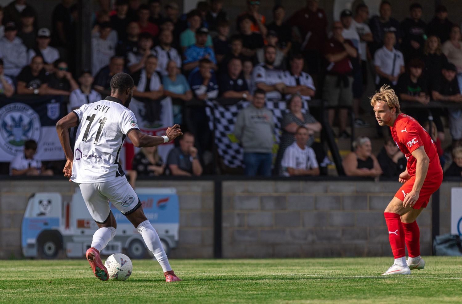 Two soccer players compete for the ball during a sunny outdoor match with a crowd of spectators in the background.