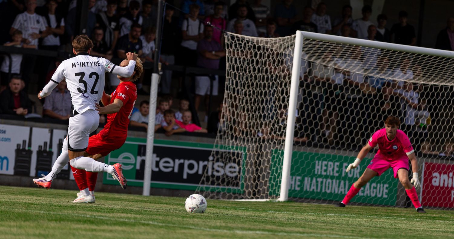 Player number 32 shoots on goal during a competitive football match, with goalkeeper in green ready to defend.