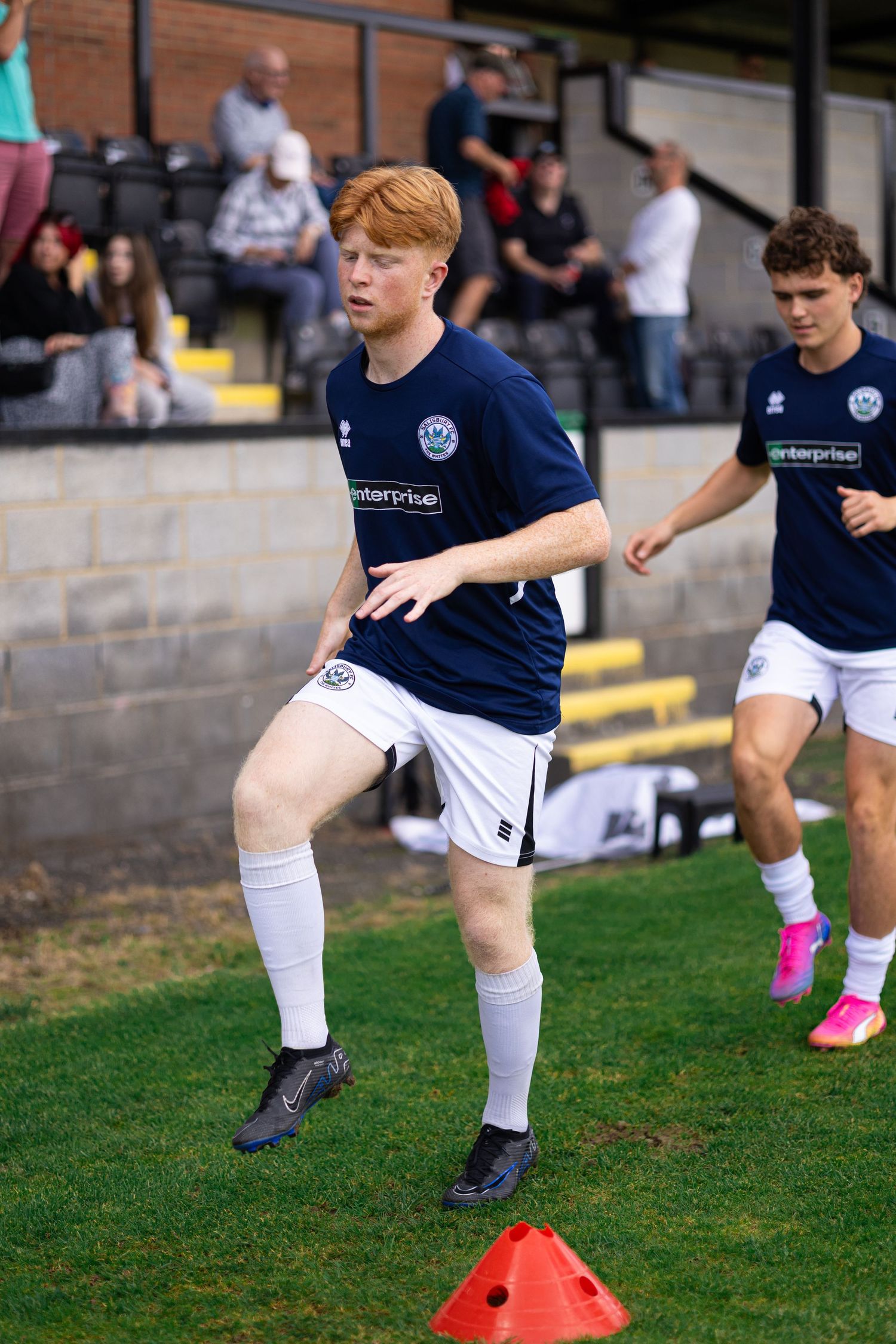 Young redhead soccer player in navy kit warming up with cones on pitch, fans visible in stands behind.