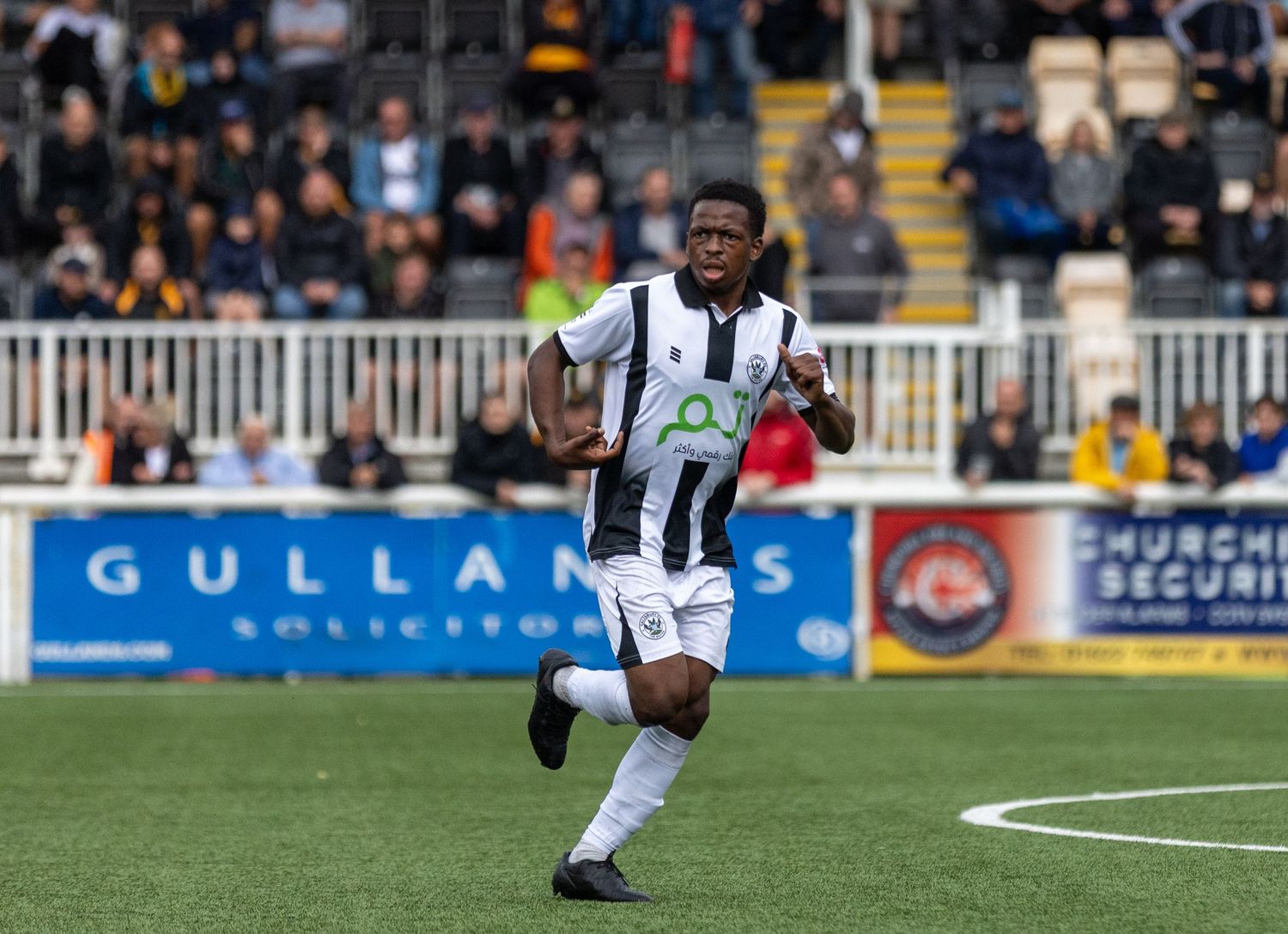 A footballer in black and white kit sprints on the pitch during a match at a stadium with advertising boards.