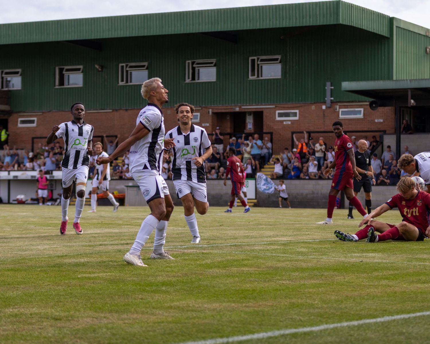 Football players in white celebrate a goal together on the pitch during a sunny outdoor match.