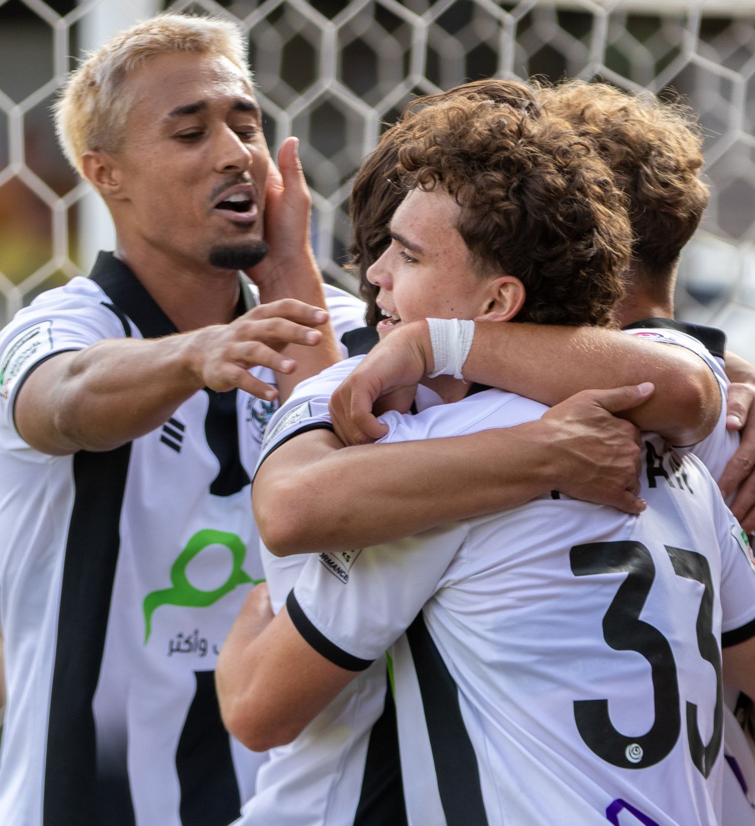 Footballers in white kits celebrate together on the pitch, with player number 33 receiving congratulations from teammates.