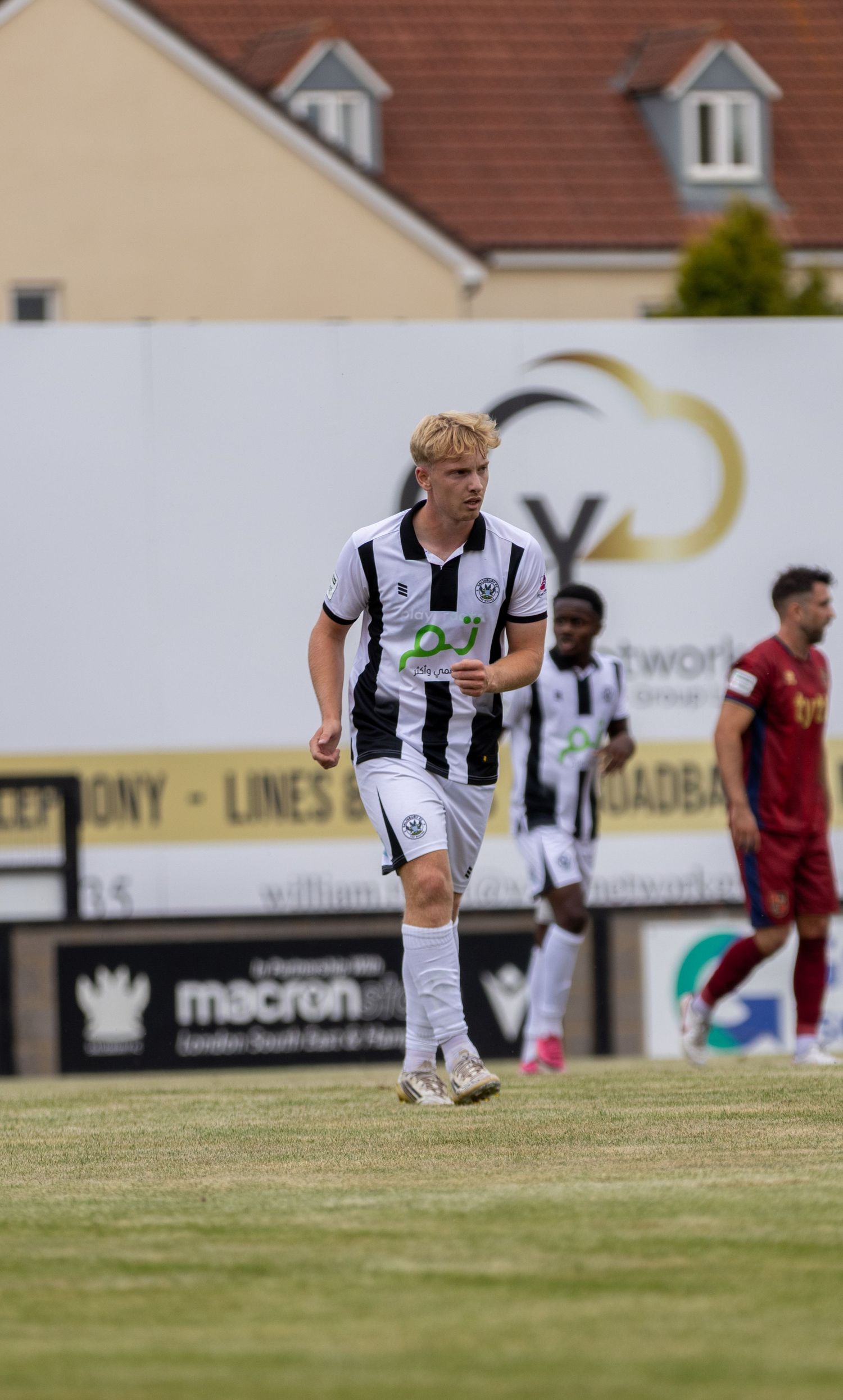 Blonde footballer in black and white striped kit jogs during outdoor football match daytime.