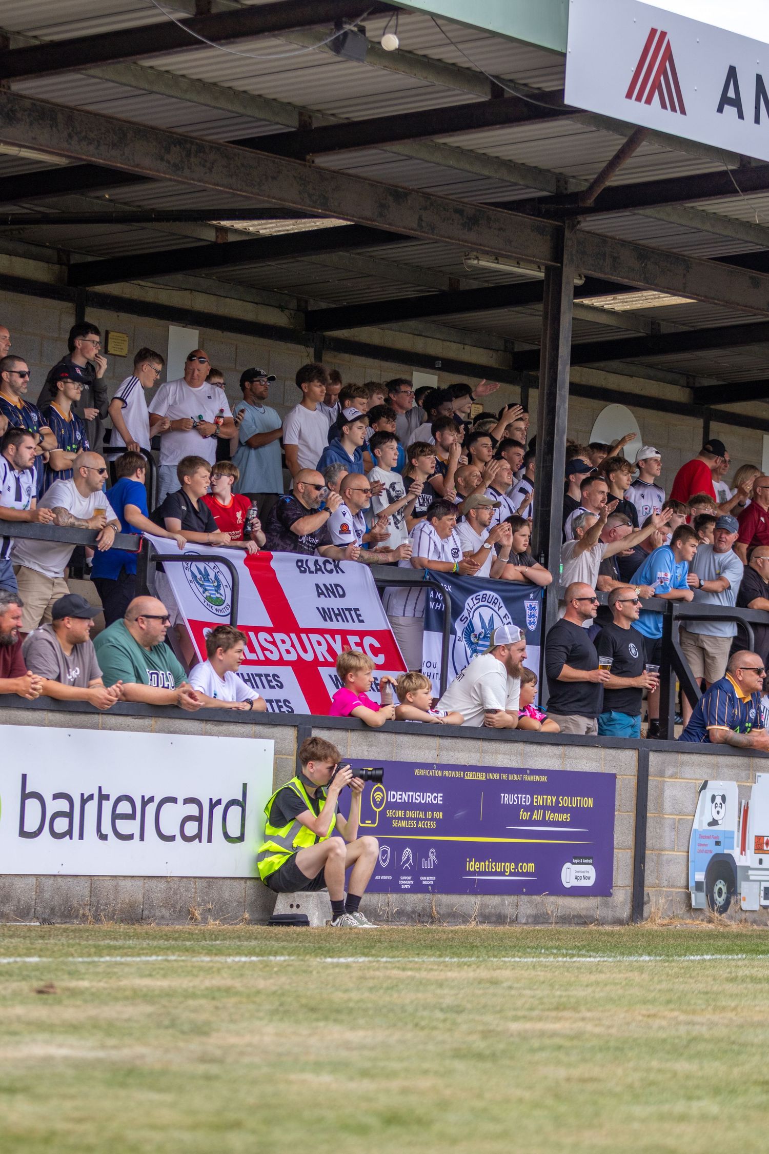 Salisbury FC fans display a Black and White banner in a packed stand during a sunny home league match.