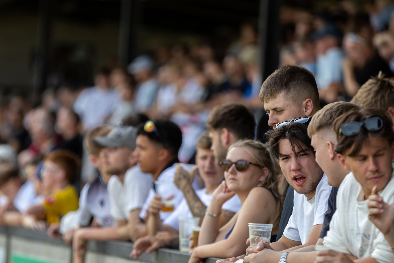 Young football fans lean on a railing watching a match from the stands on a sunny day.