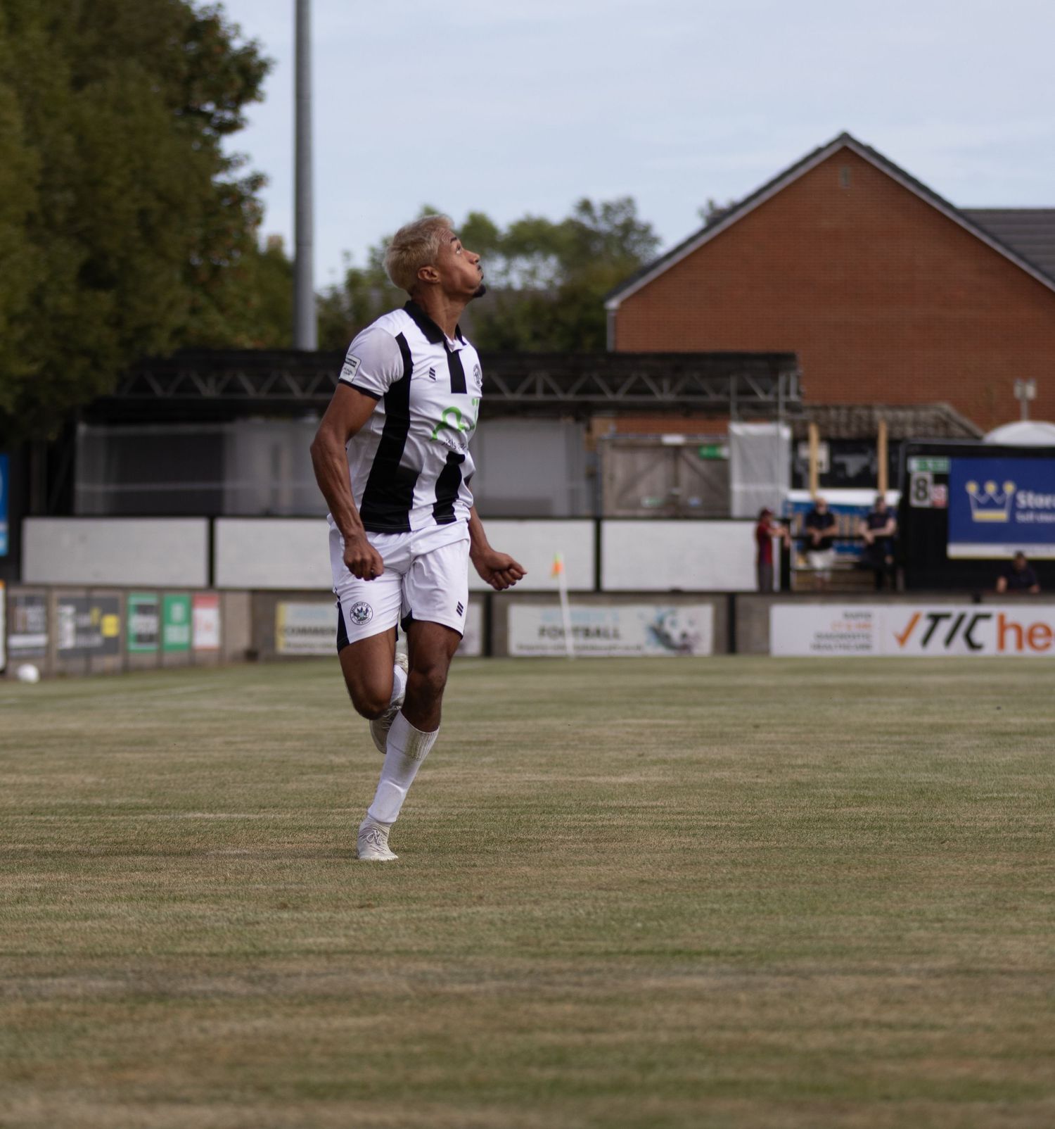 Soccer player in black and white striped kit celebrates on grass pitch with stadium stands visible in background.