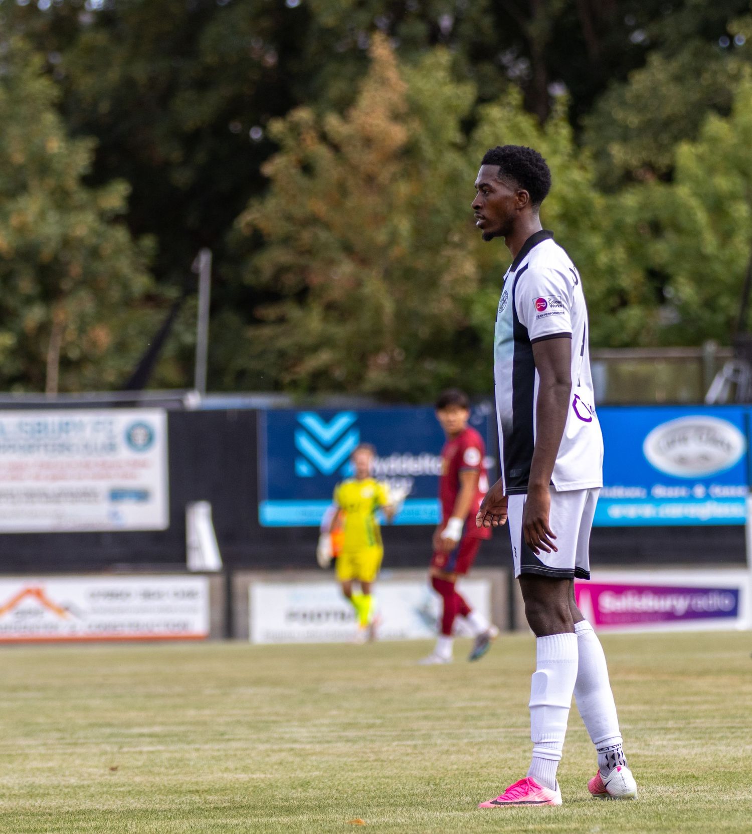 Football player in white kit stands on pitch with opposition in red visible in the background.