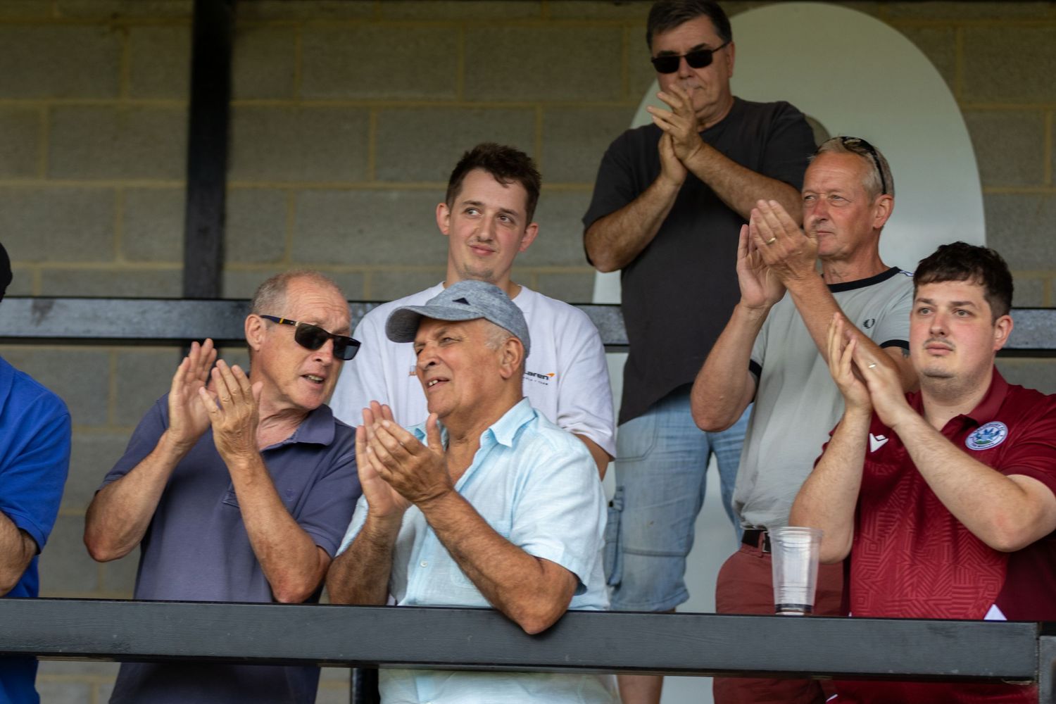 Spectators applauding in a covered stand at a football match on a sunny day.