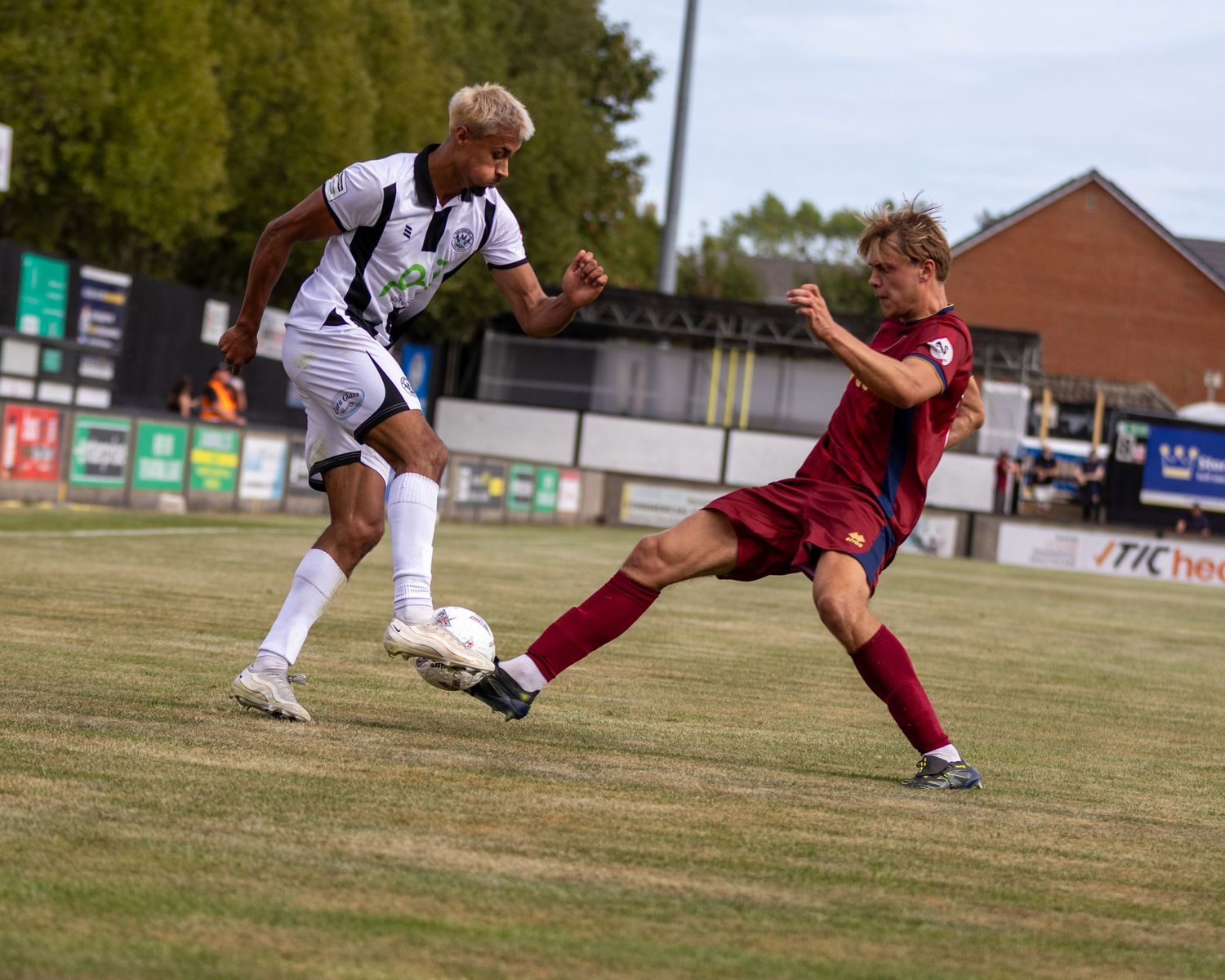 Two soccer players compete for ball, white kit versus red kit, during match on green grass pitch.