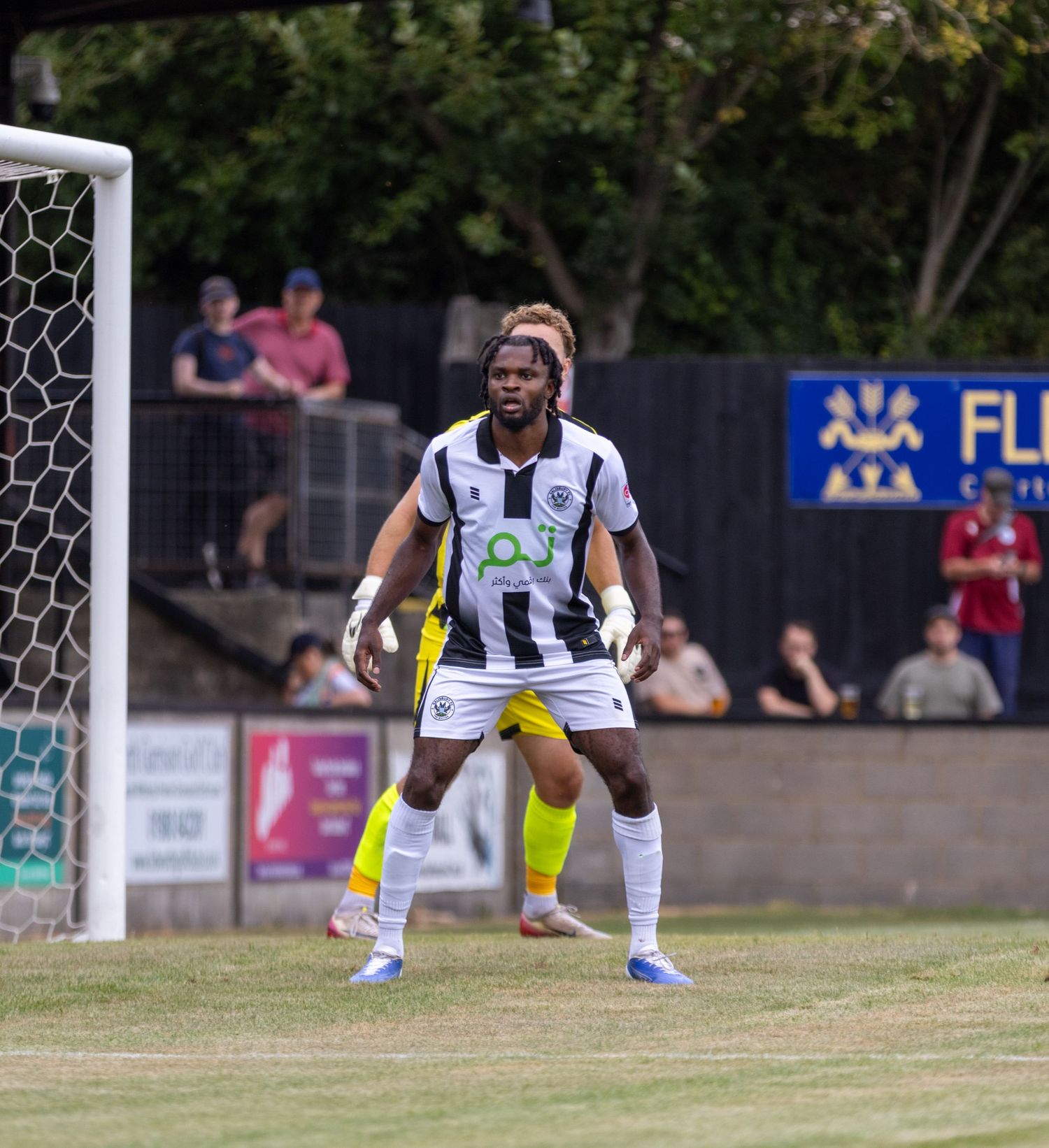 A Black footballer in black and white striped kit stands alert near the goal post.