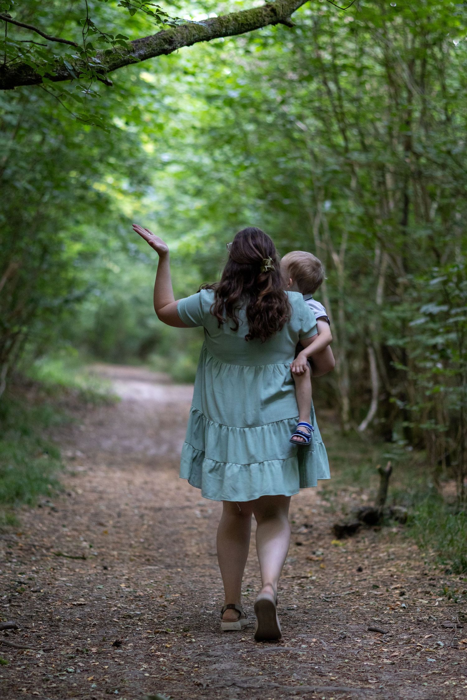 Mother carrying toddler while walking along a green forest path, waving overhead.
