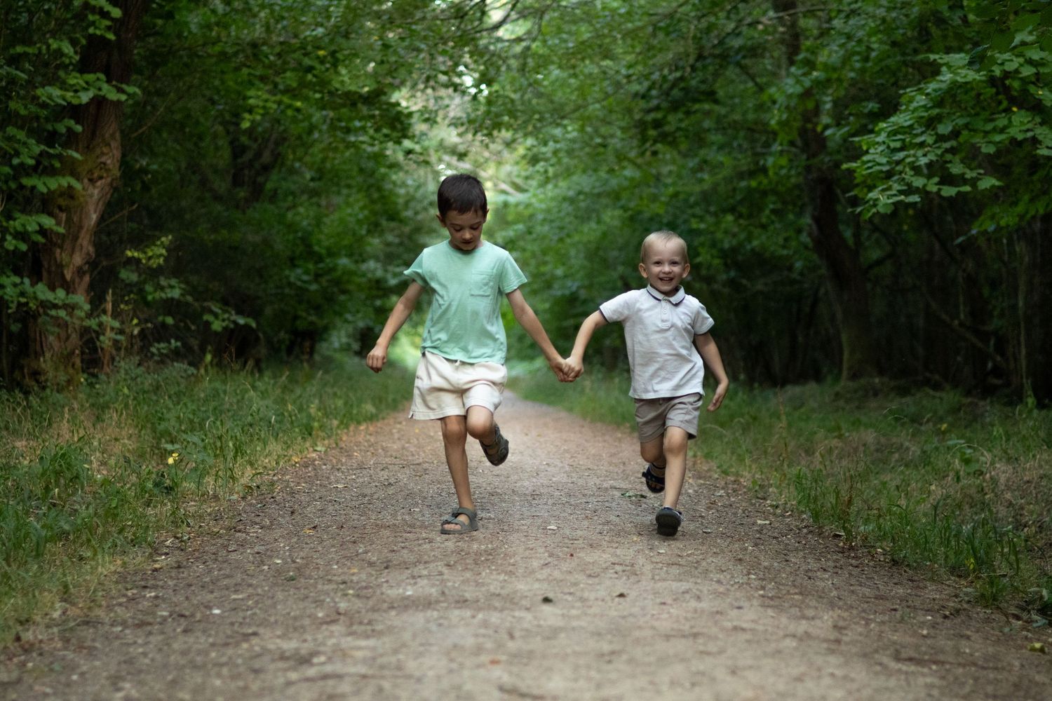 Two children holding hands running together on a forest dirt path surrounded by lush green trees.