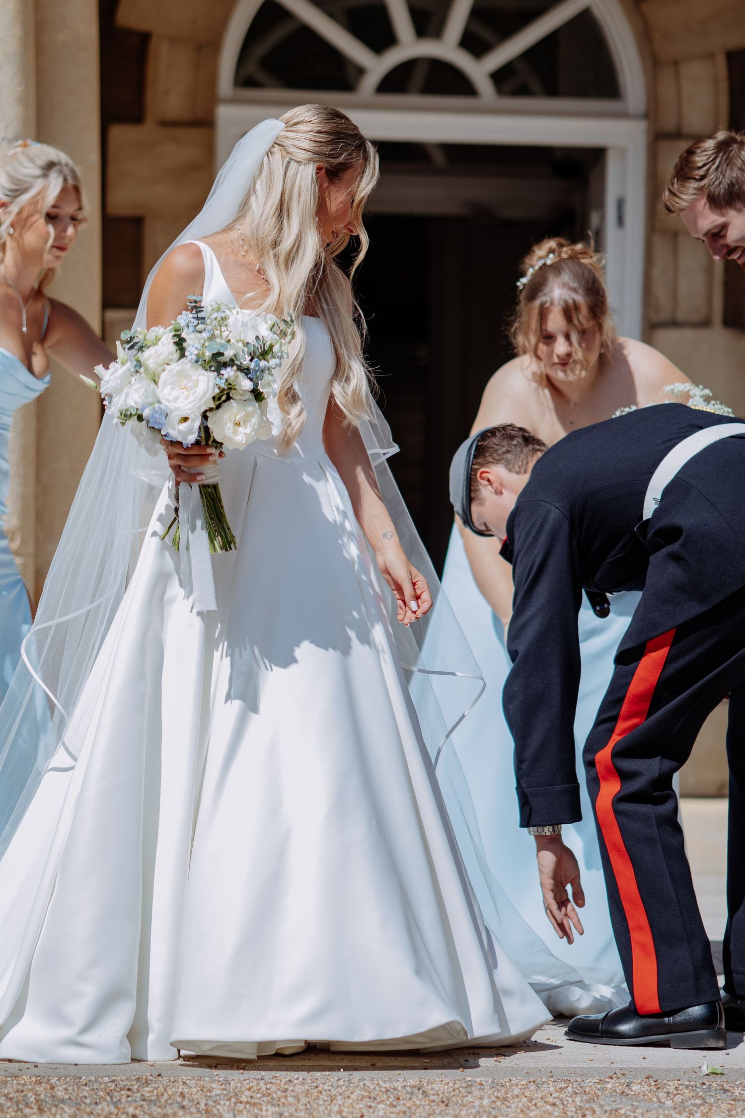Groom in military uniform fixes bride's dress with bridesmaids assisting outside.