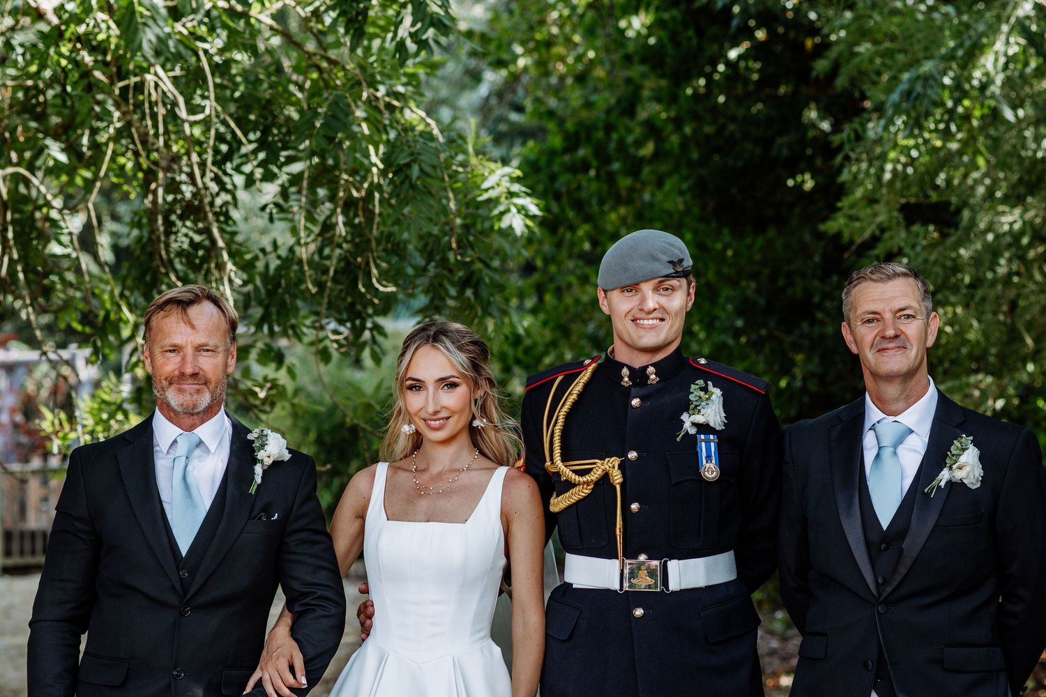 Wedding group photo with bride, groom in military uniform, and two men in suits outdoors.