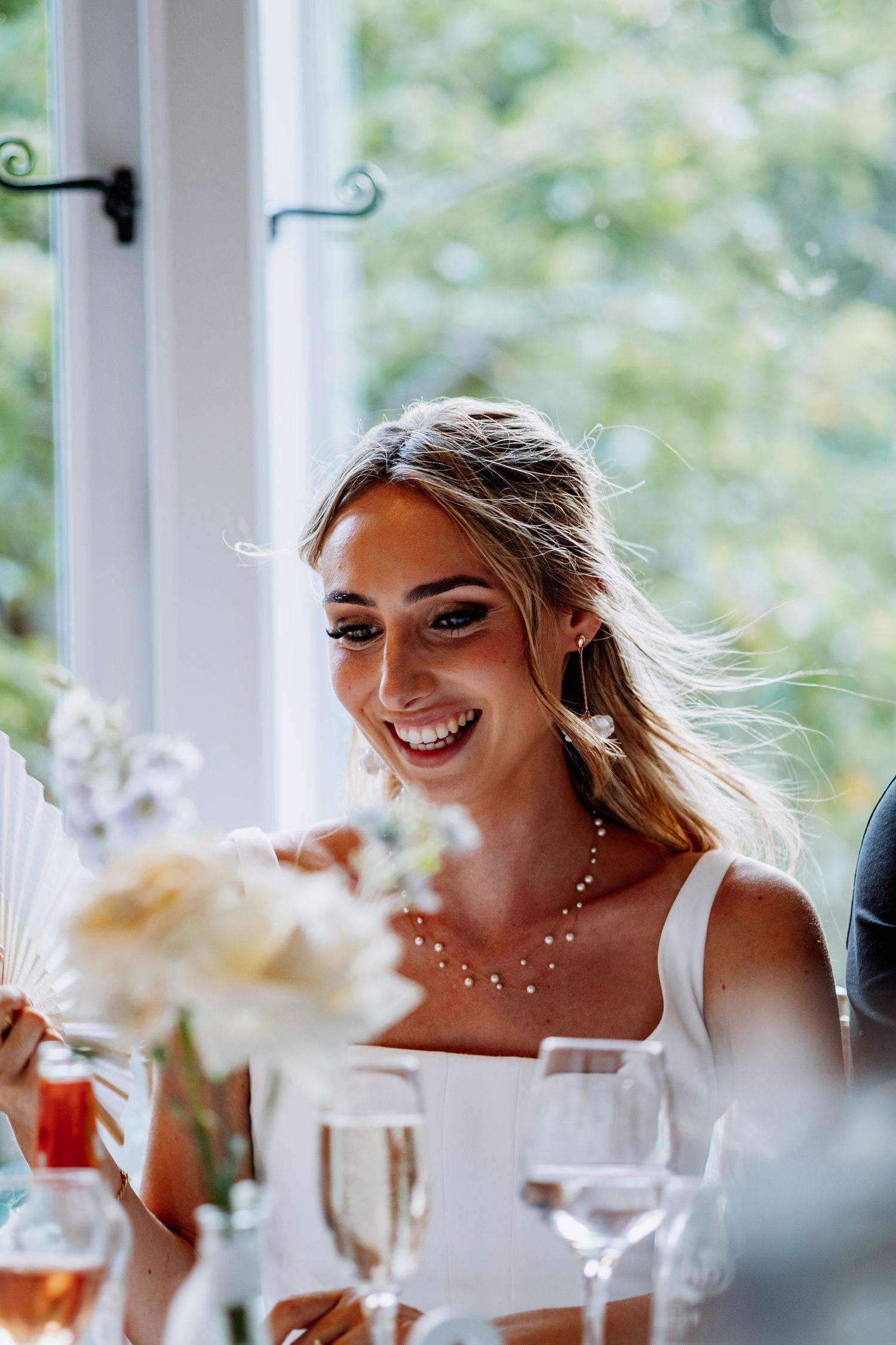 Radiant blonde bride in white dress laughing at wedding reception table surrounded by champagne glasses and flowers.