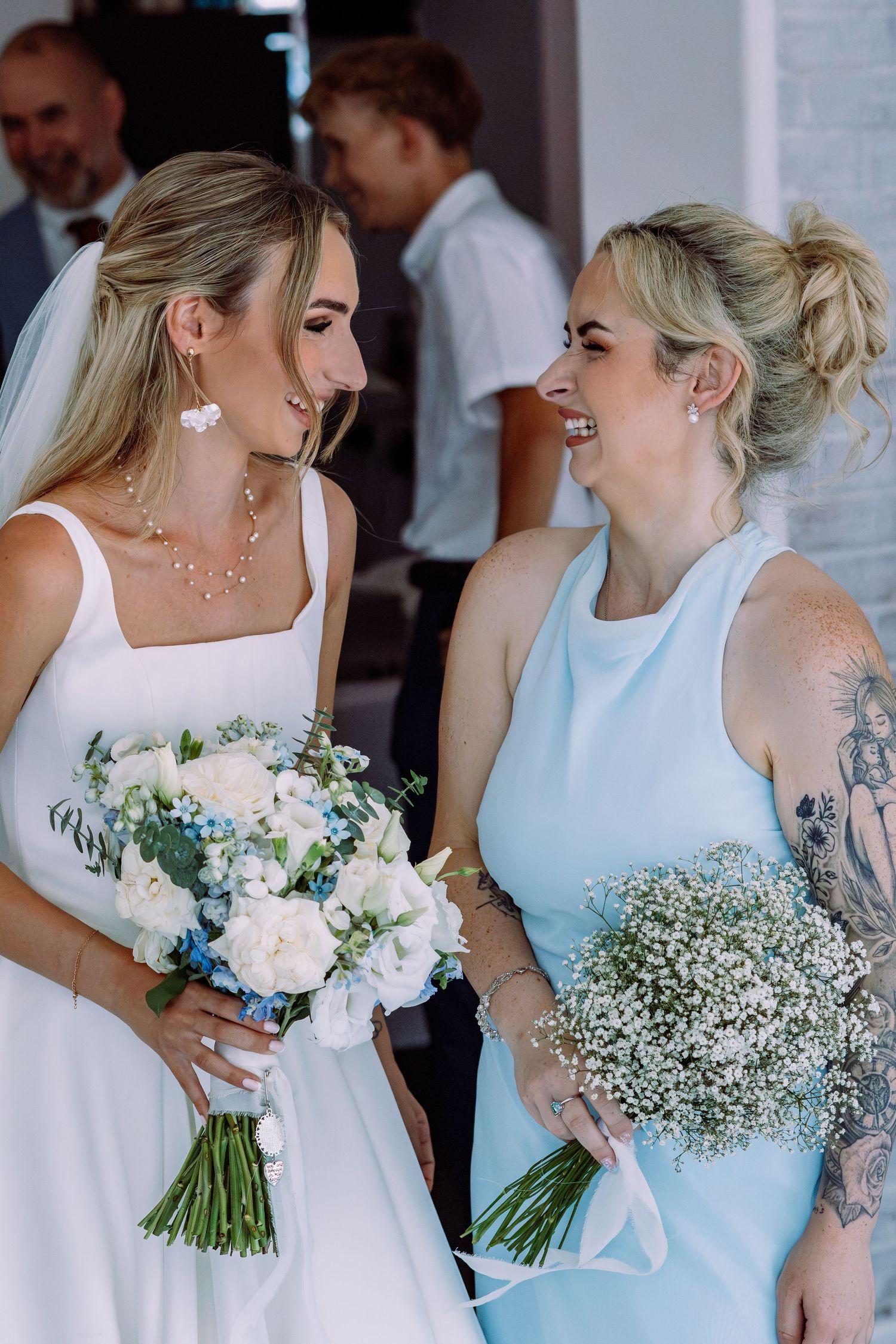 Bride and tattooed bridesmaid in blue dress laughing together while holding wedding bouquets.