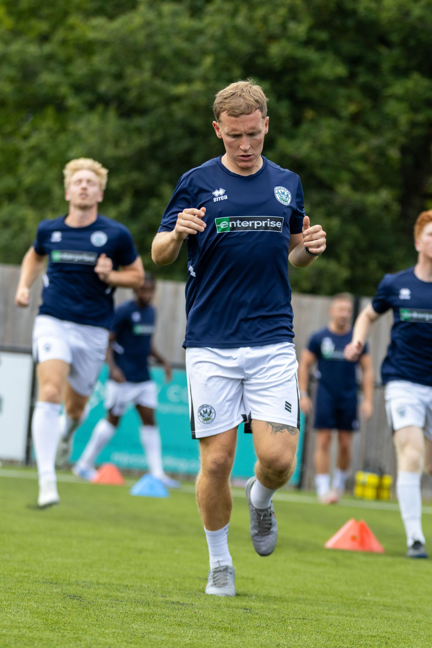 Football player in navy Enterprise kit leads group of teammates during pre-match running warmup on grass pitch.