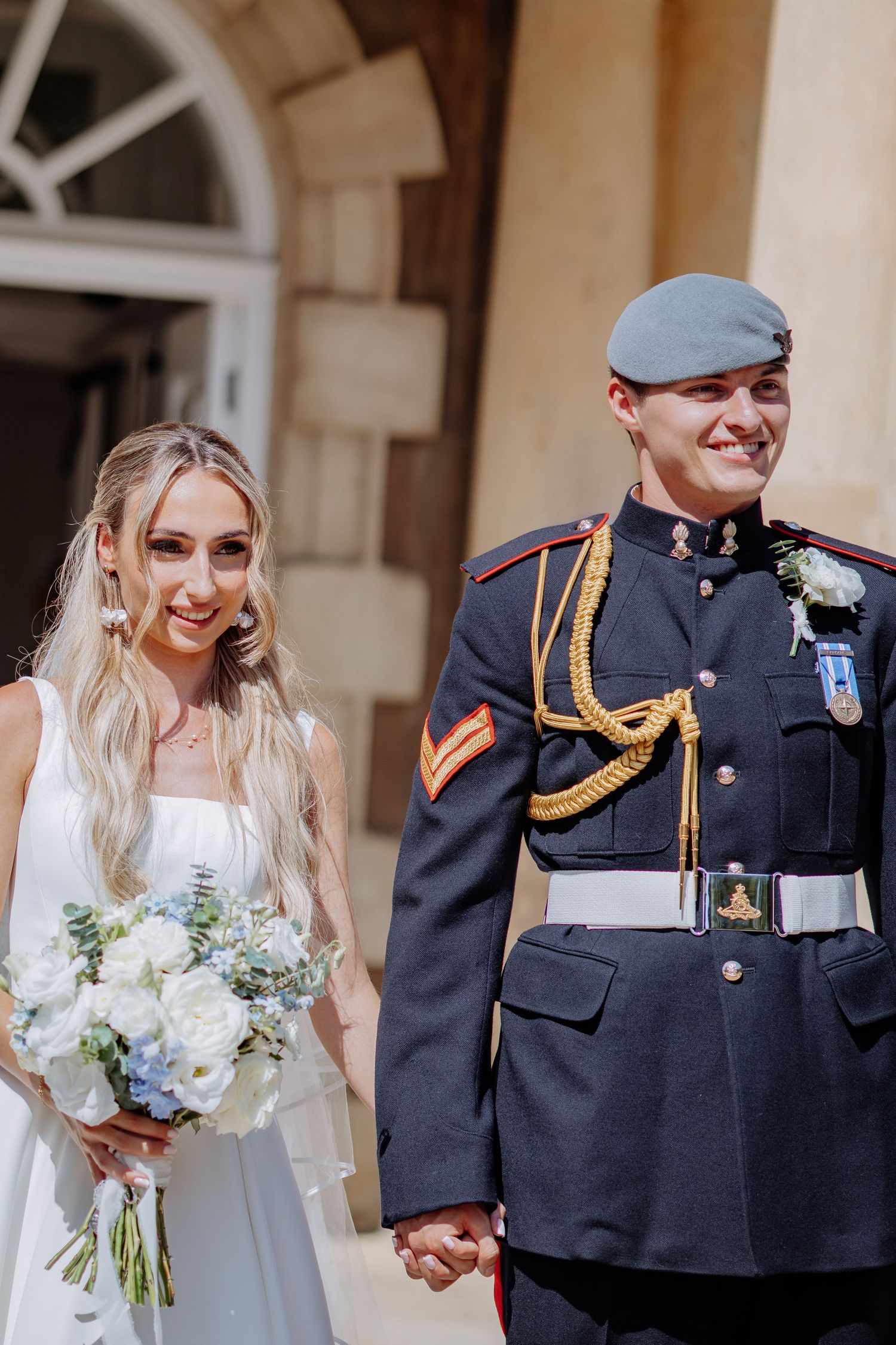 Smiling bride in white gown holds blue and white bouquet beside groom in RAF ceremonial uniform with gold aiguillette.