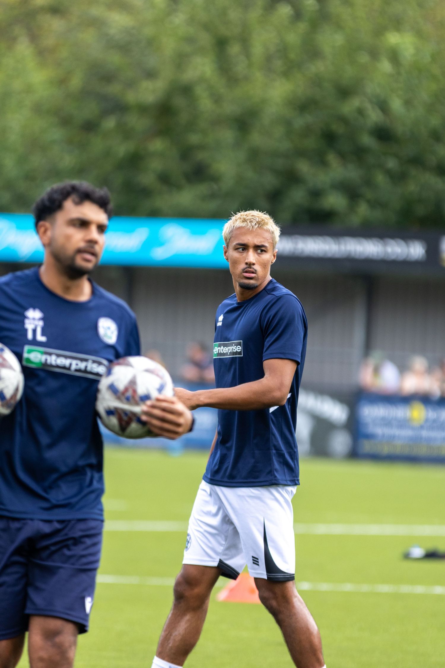 Two footballers in navy Enterprise-sponsored kits handle ball during pre-match warmup session outdoors.