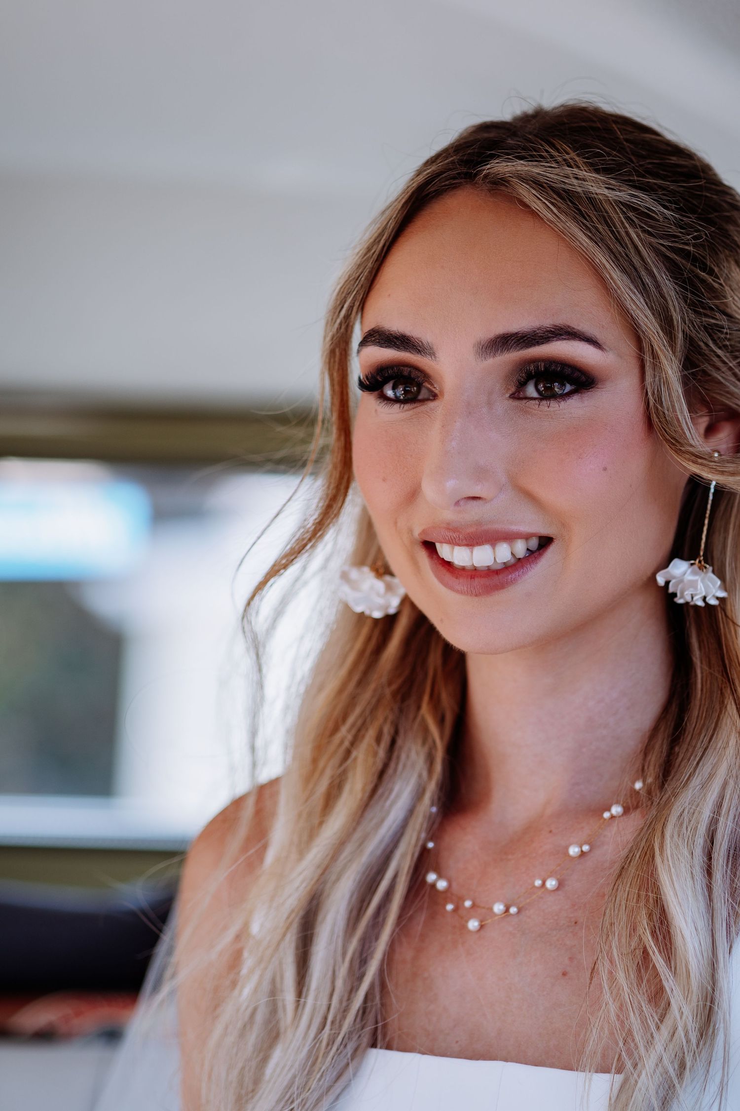 Beautiful bride smiling warmly wearing white floral drop earrings and delicate pearl necklace on wedding day.