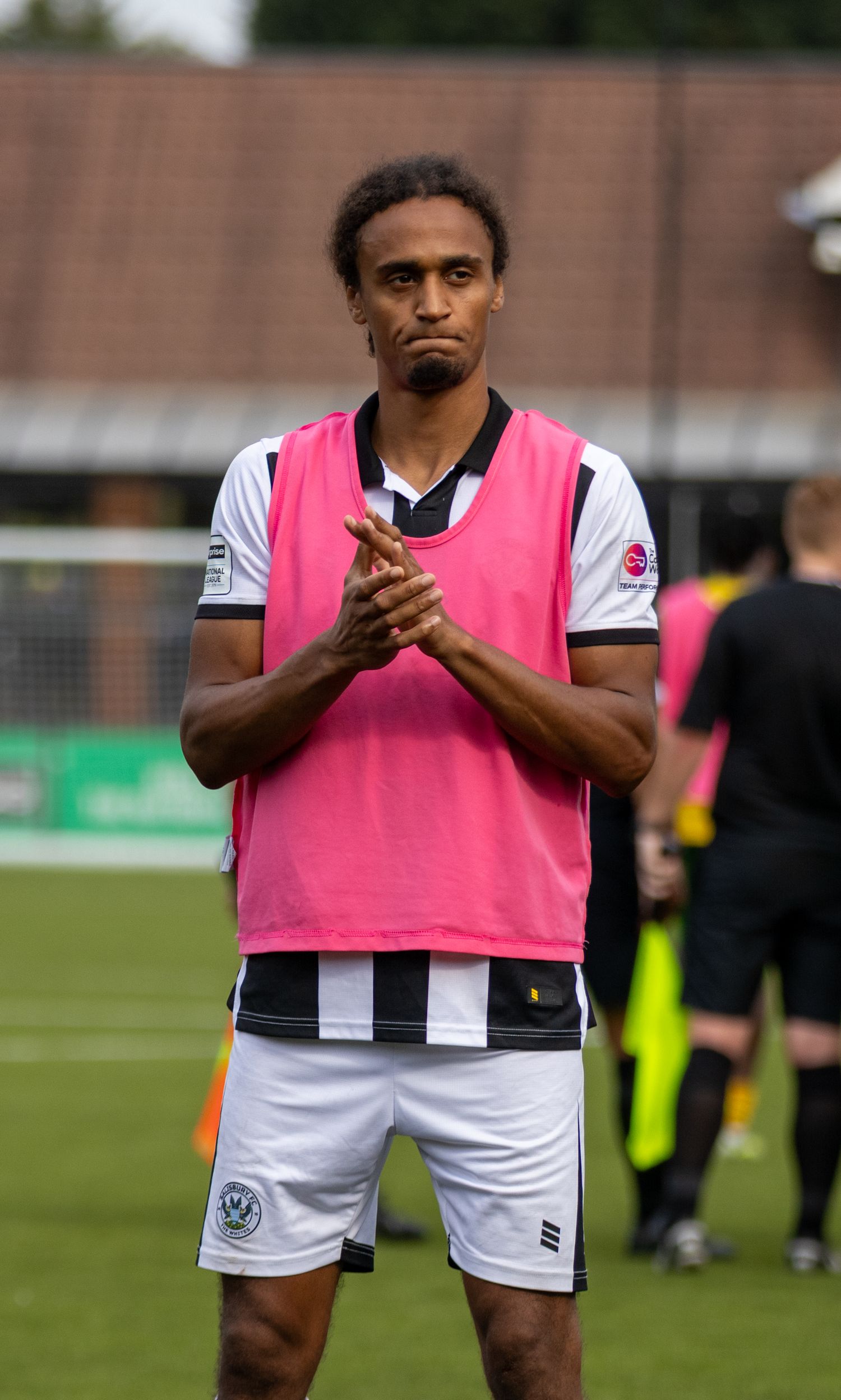 A footballer in black and white striped kit with pink bib applauds fans on a football pitch.
