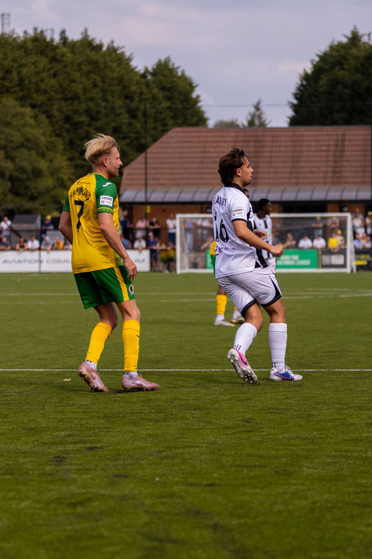 Yellow-green and white-kitted footballers position themselves during a break in play on a sunny football pitch.